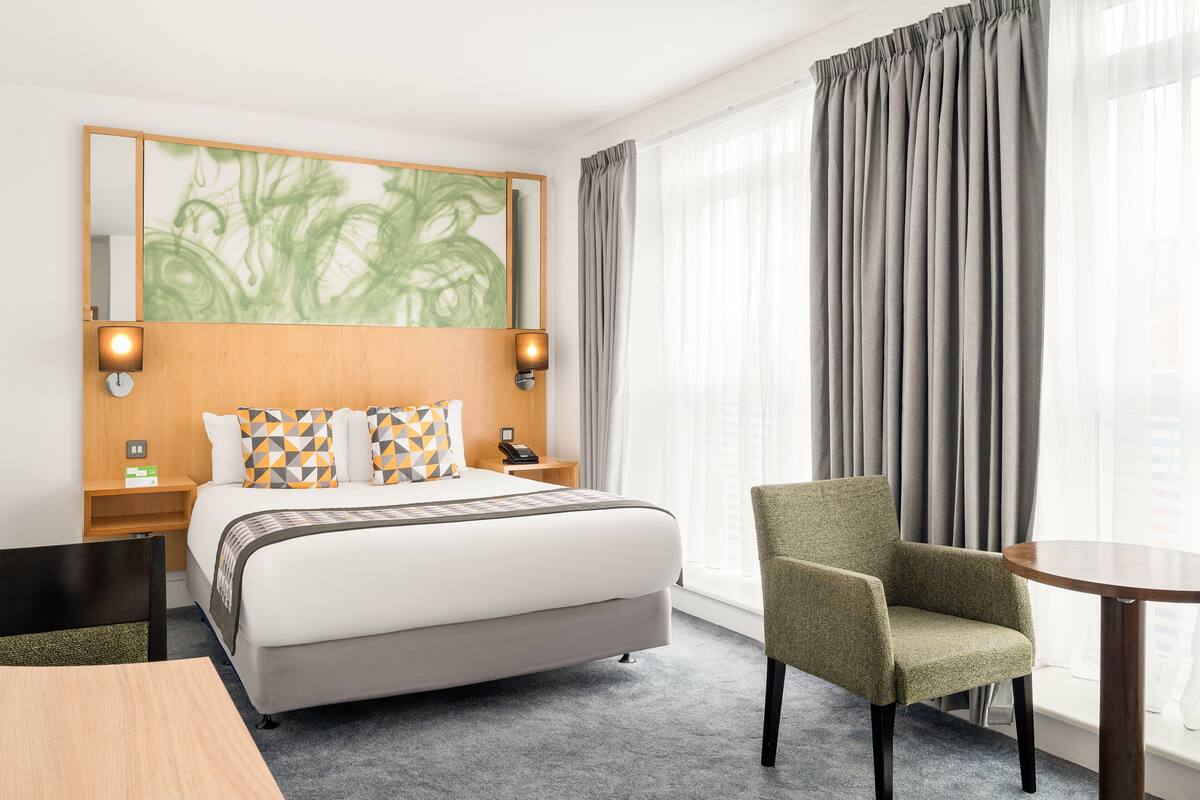 A hotel room with a large bed, two bedside tables, a chair, a small table, and a large window with curtains.