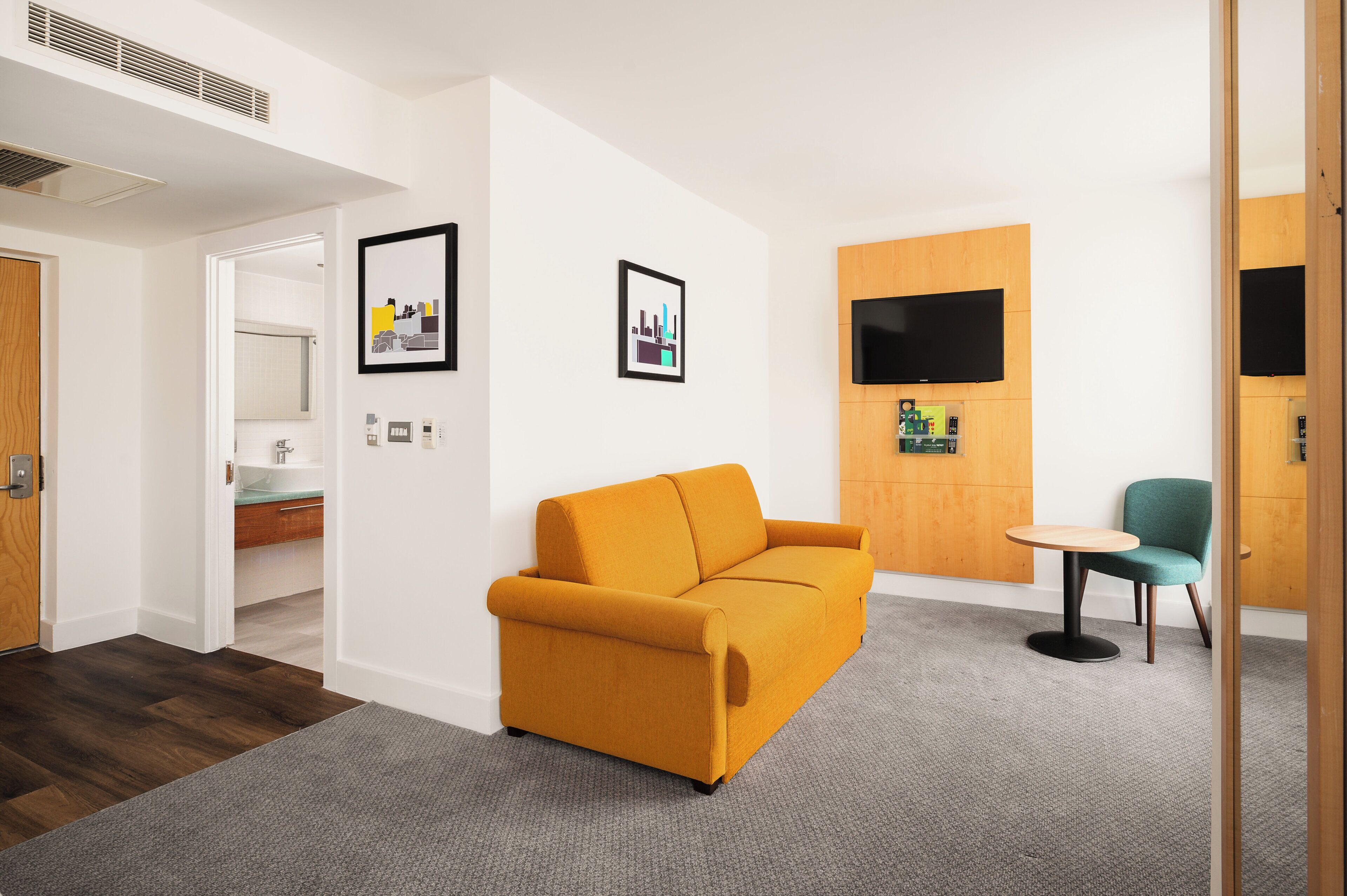 A modern hotel room with a mustard sofa, a teal armchair, a small round table, and a flat-screen TV mounted on a wooden panel.