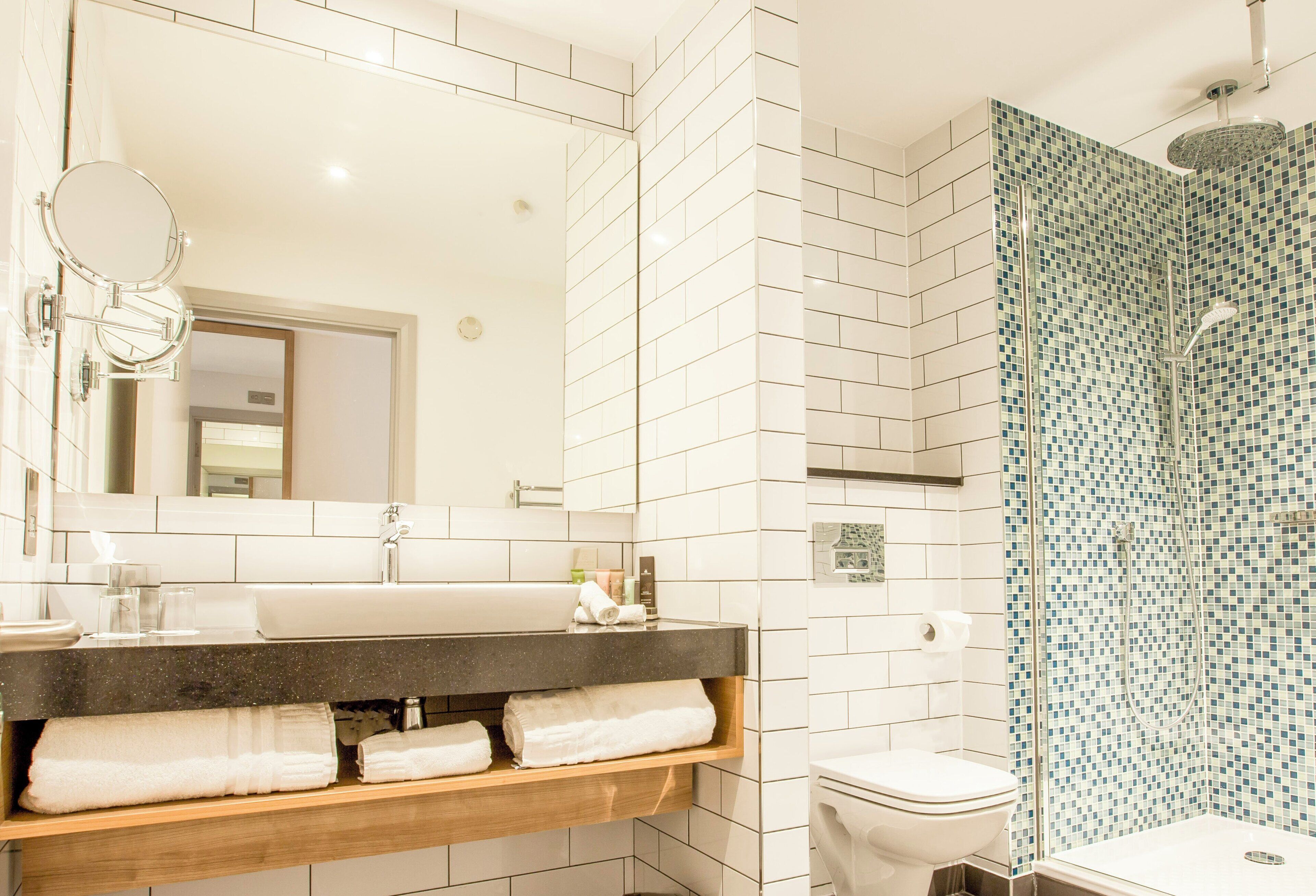 A modern bathroom with a large mirror, white tiles, a shower with a glass door, and a wooden vanity with towels.