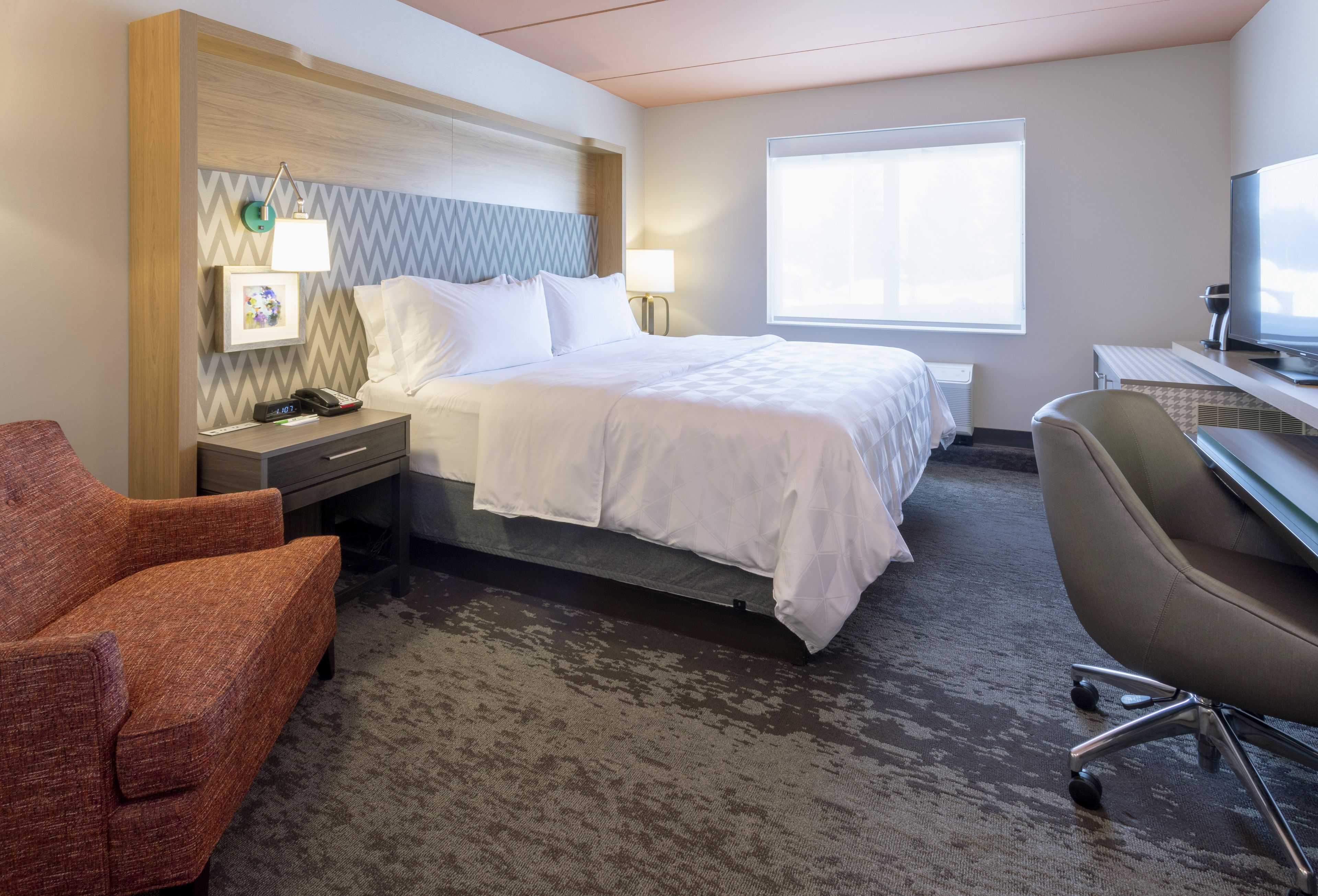 A hotel room with a bed, a desk with a computer, a chair, a nightstand, and a window.