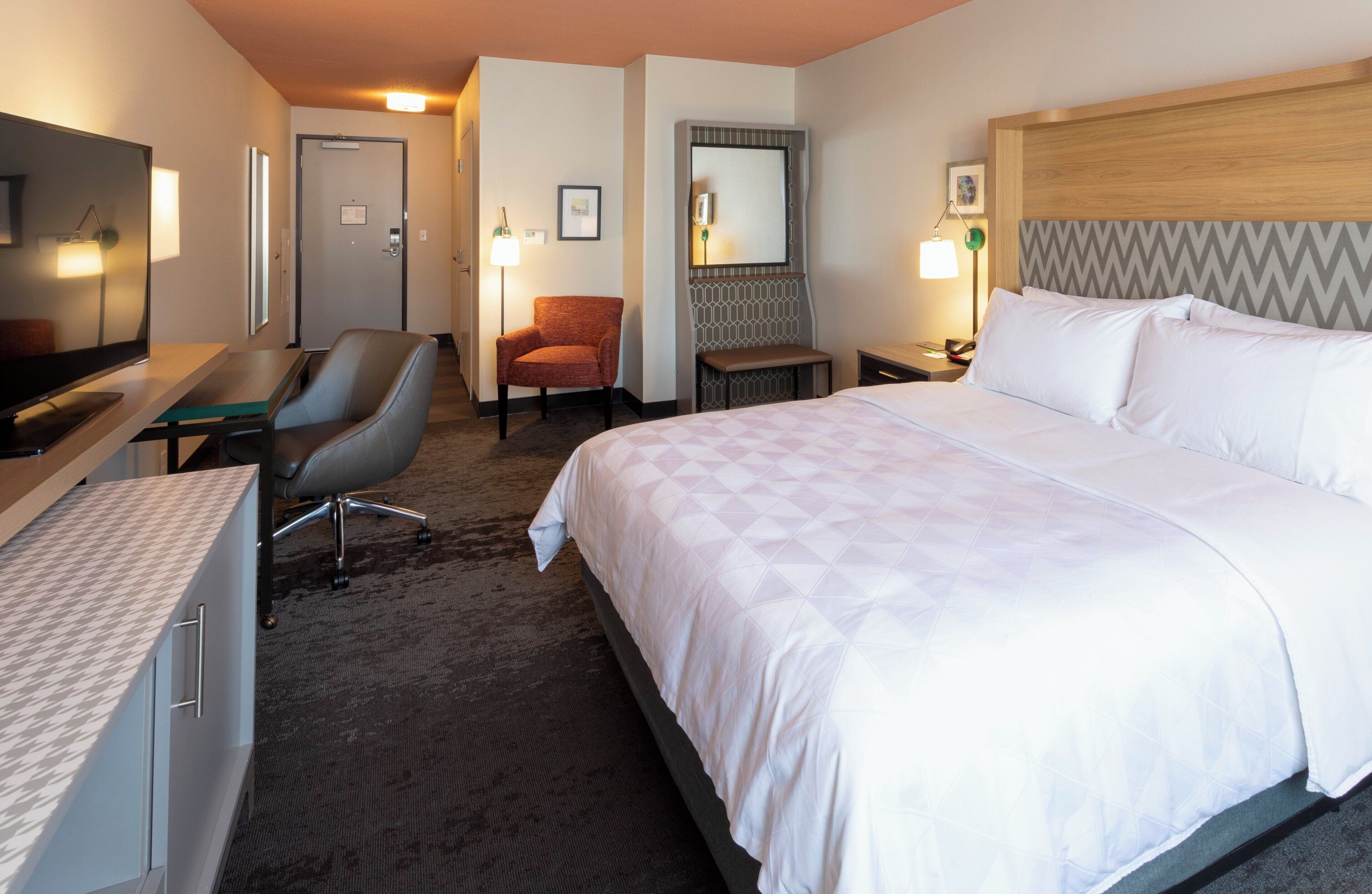 Habitación de hotel con cama, escritorio, silla, televisor y espejo.