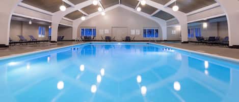 Una piscina interior con agua cristalina, rodeada de sillas reclinables y mesas, bajo un techo alto con vigas de madera.