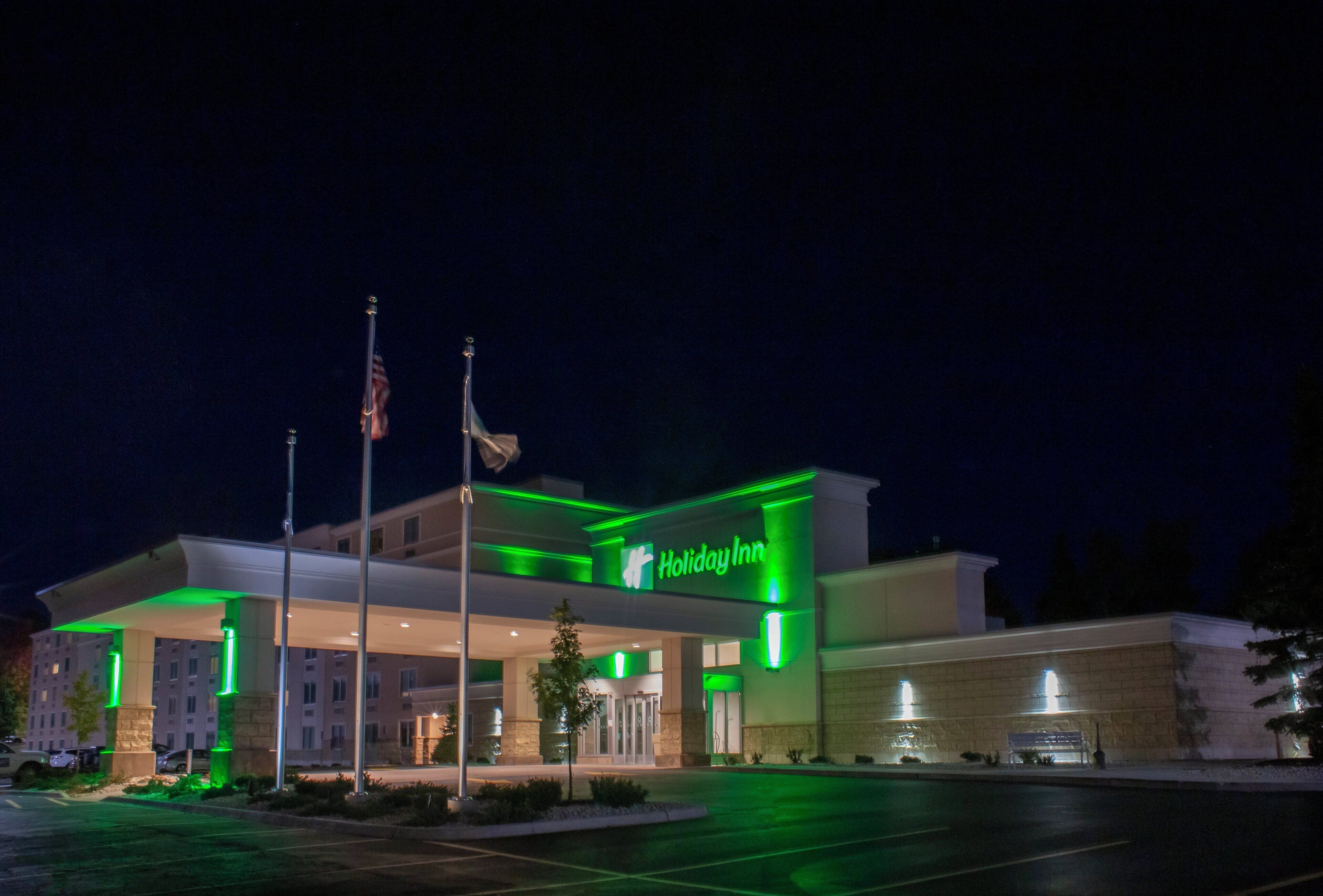 A Holiday Inn hotel with a modern design, featuring a prominent green sign, a flagpole with flags, and a well-lit entrance.