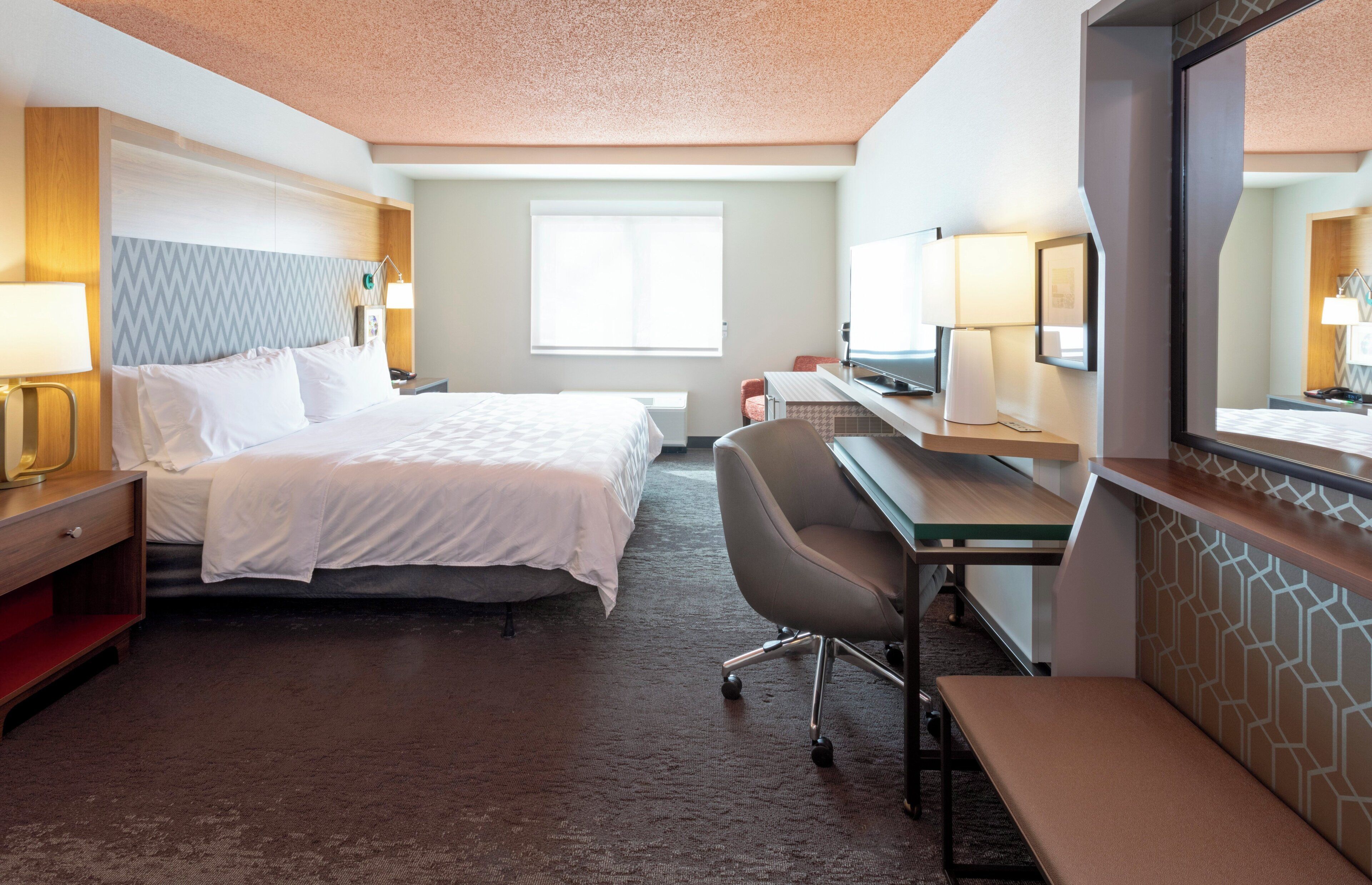 A hotel room with a bed, desk, chair, and a large mirror.
