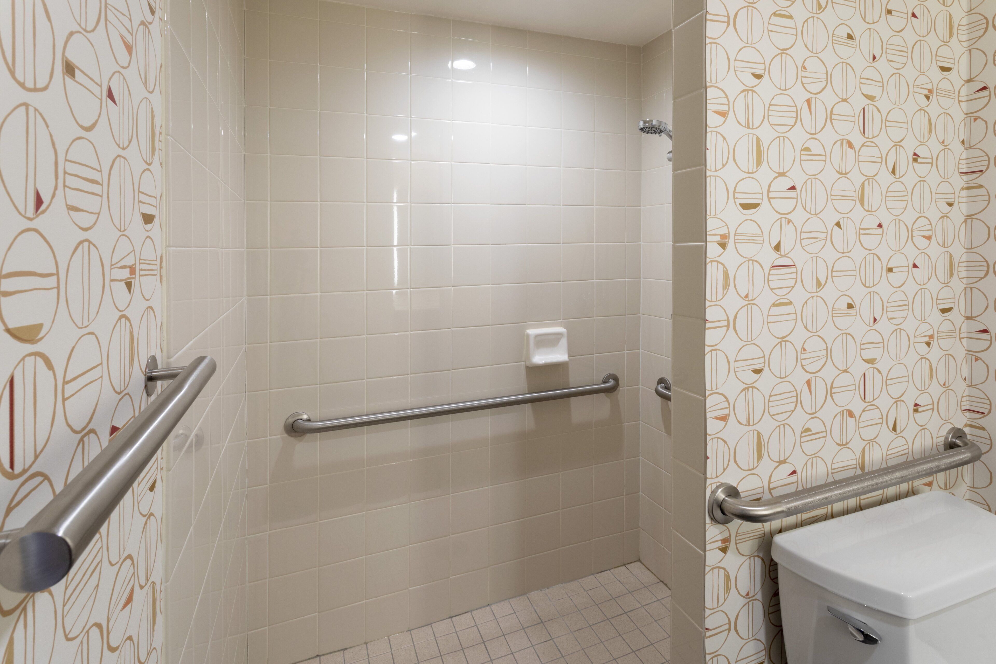 Une salle de bain avec des murs recouverts de carreaux blancs, des toilettes et une barre d’appui.