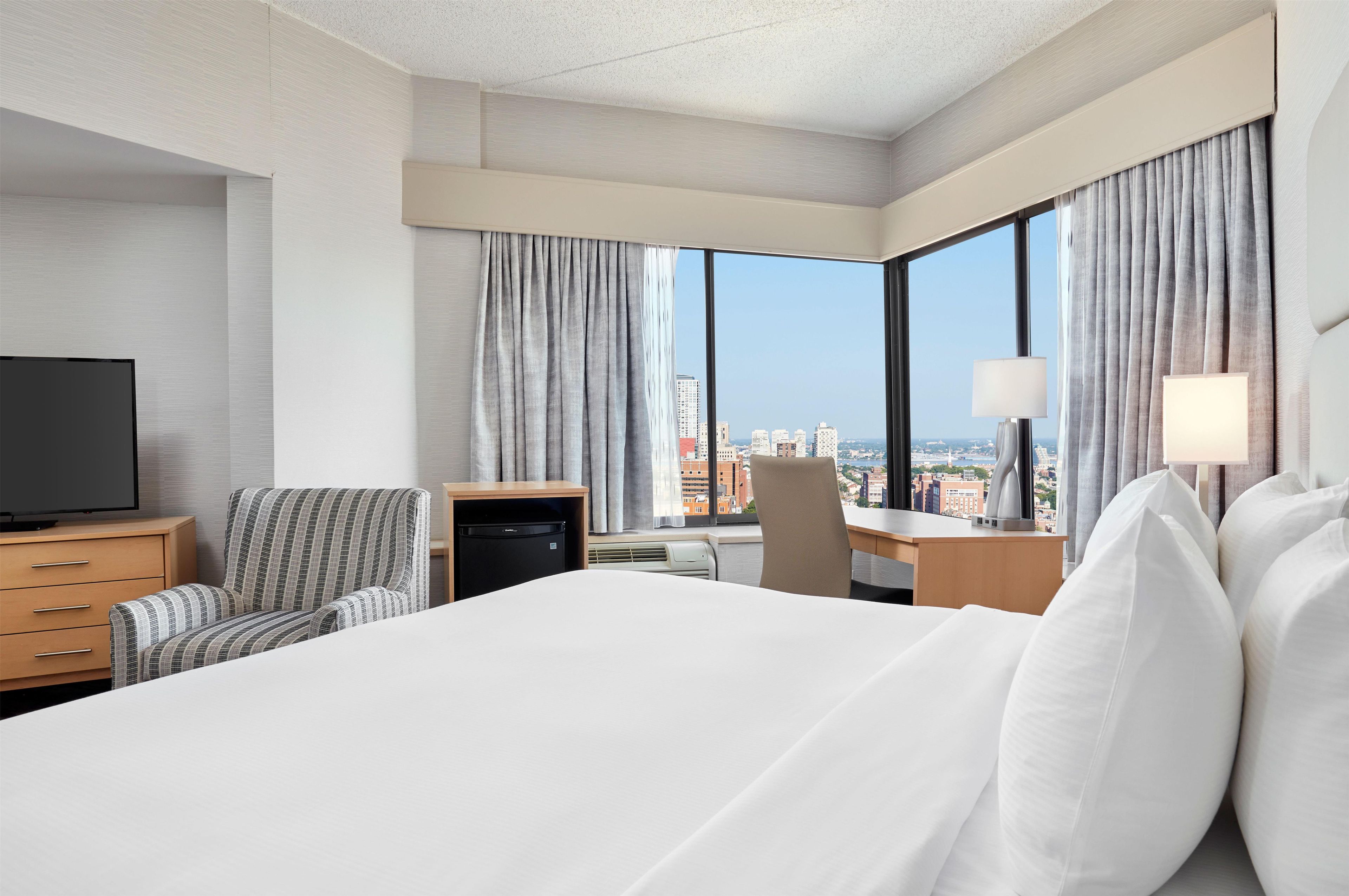 Habitación de hotel con una cama grande, un escritorio, una silla, un televisor y vistas a la ciudad.