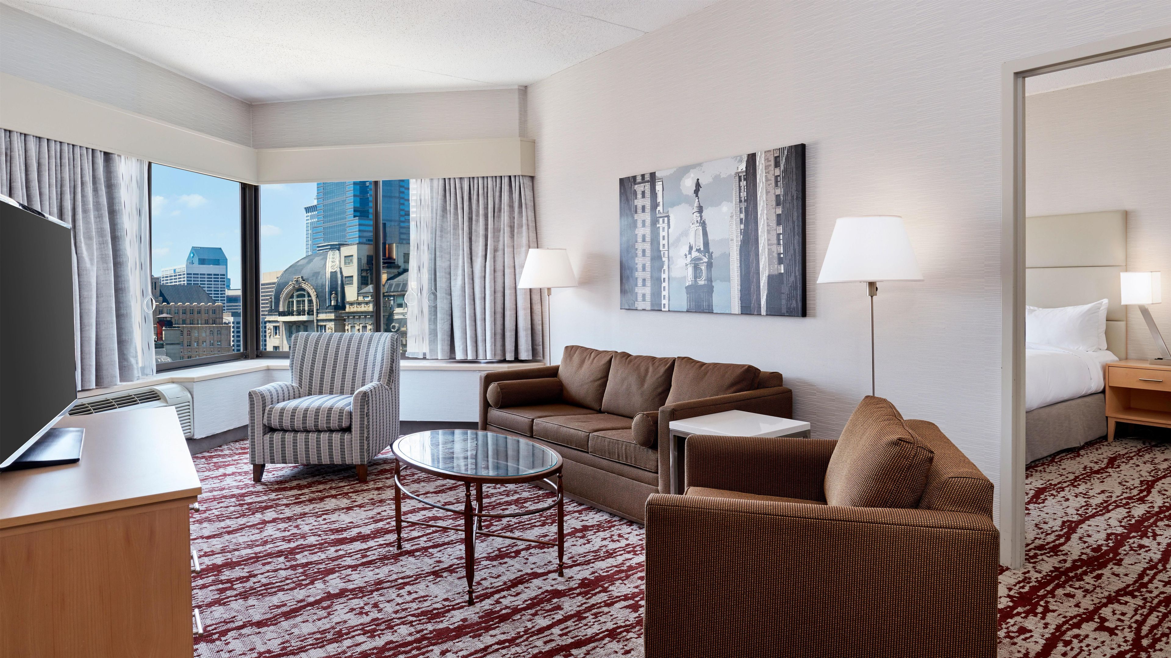 A hotel room with a large window, a sofa, a chair, a small table, and a TV.