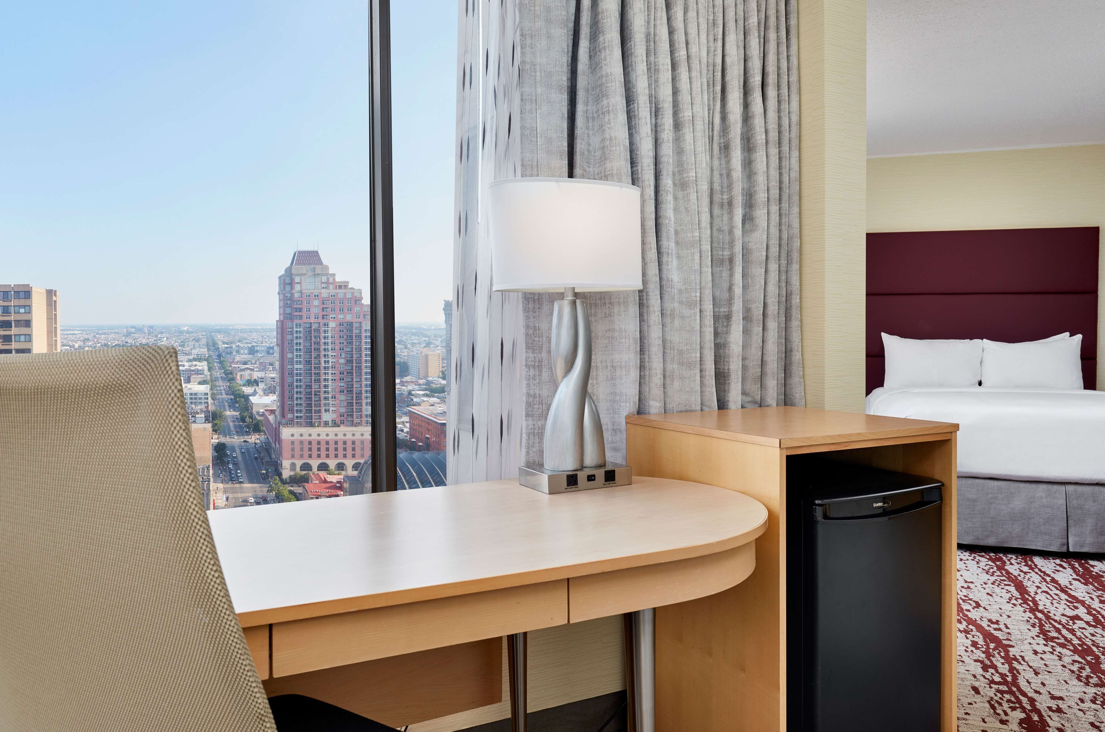 Ein Hotelzimmer mit Blick auf die Stadt, einem Schreibtisch mit Lampe, einem Bett und einem kleinen Kühlschrank.