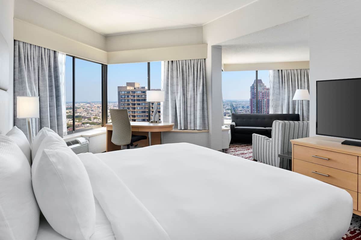A hotel room with a large bed, a desk, a chair, a TV, and a view of the cityscape.