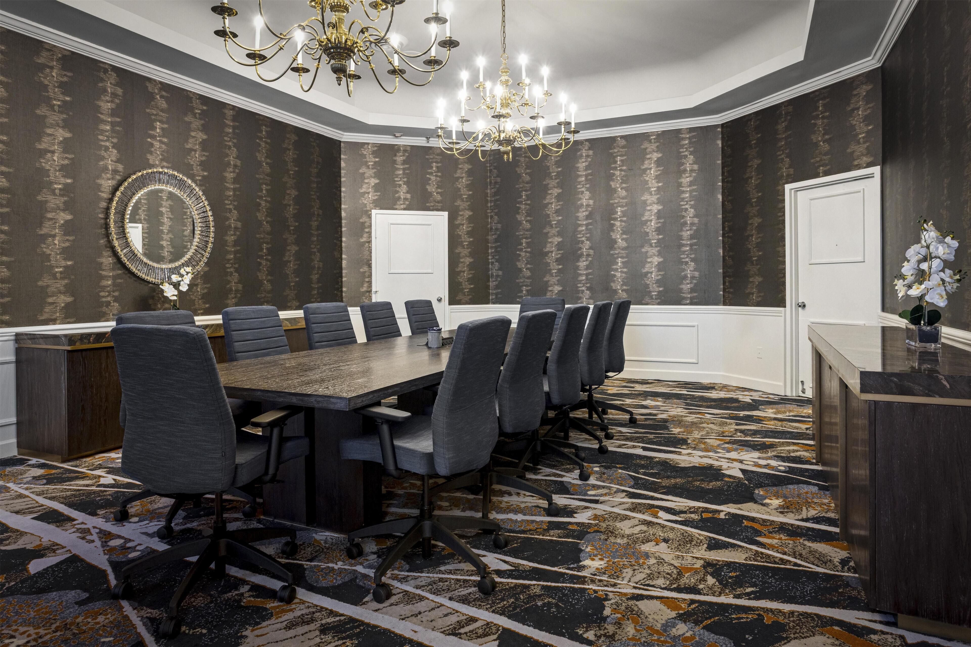 A conference room with a long table, multiple chairs, a chandelier, a mirror, and patterned carpet.
