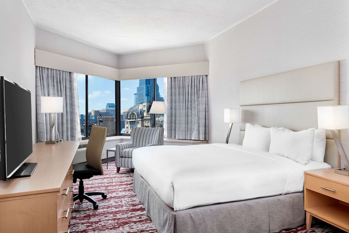 A hotel room with a large bed, a desk with a TV, a chair, and a view of a cityscape.