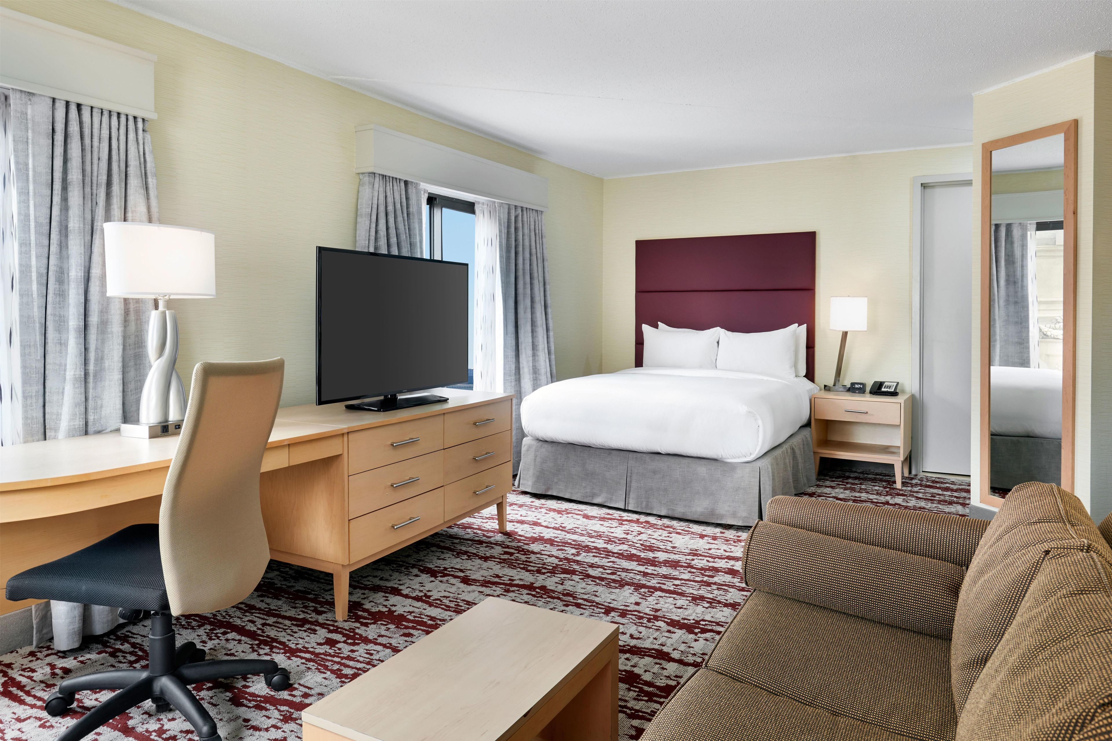 Een hotelkamer met een bed, bureau, stoel, televisie, nachtkastje en een kamerbreed tapijt met patroon.