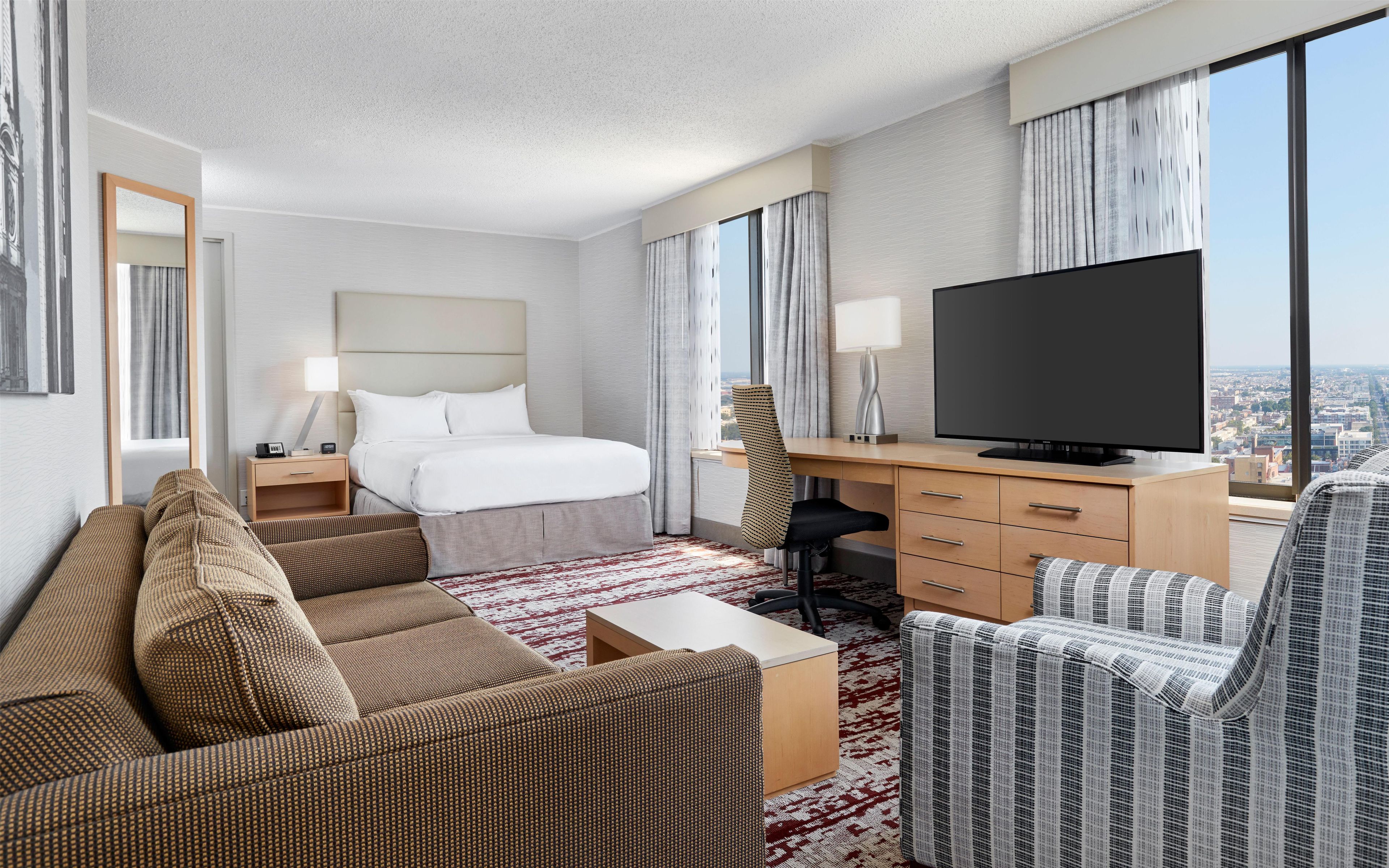 A hotel room with a bed, a desk with a TV, a sofa, and a view of the city.