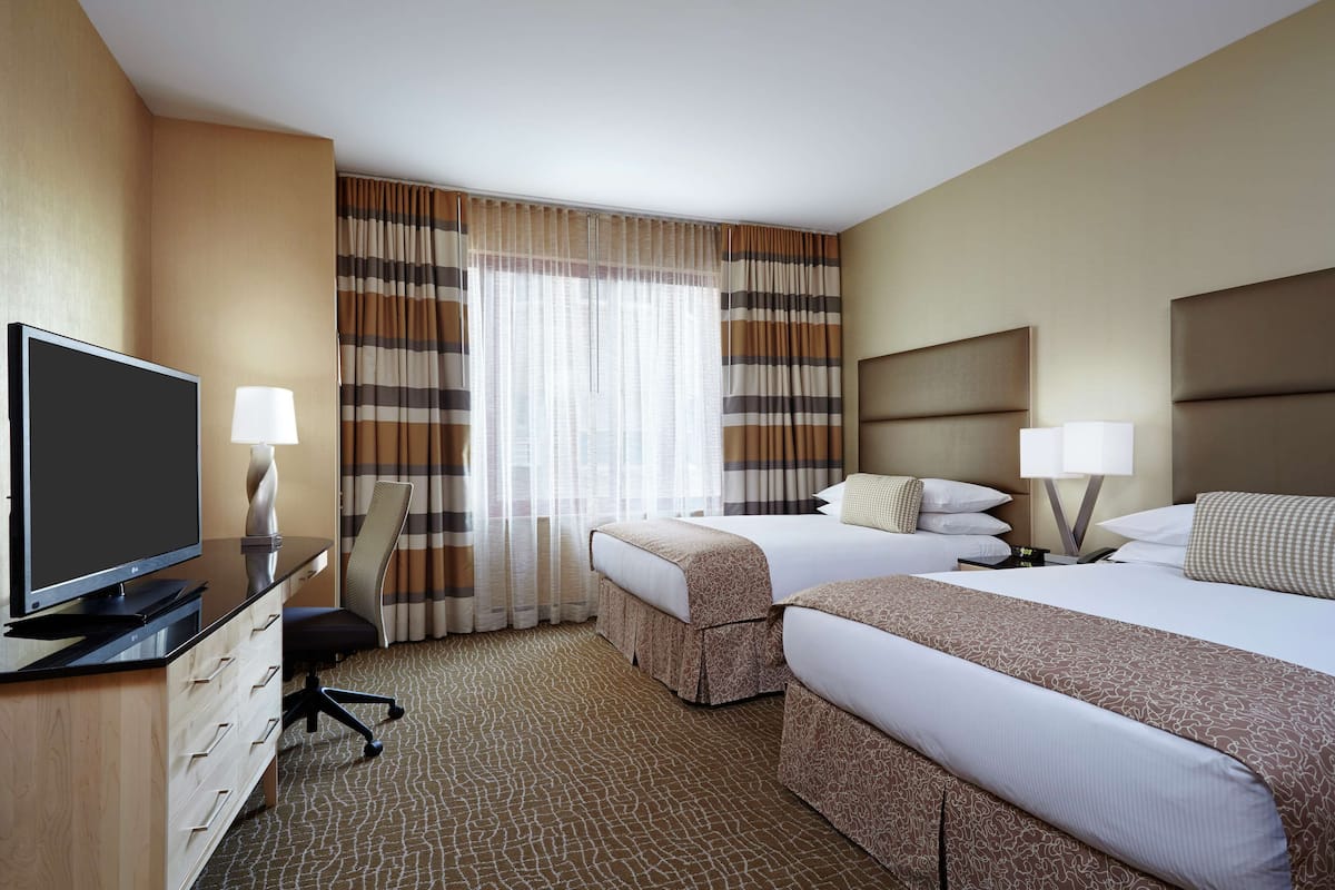 A hotel room with two beds, a desk with a television, and a large window with curtains.