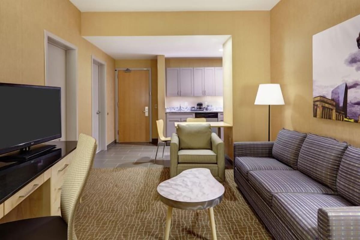 Une chambre d’hôtel équipée d’une télévision, d’un canapé, d’une petite table, d’une chaise et d’un coin cuisine à l’arrière-plan.