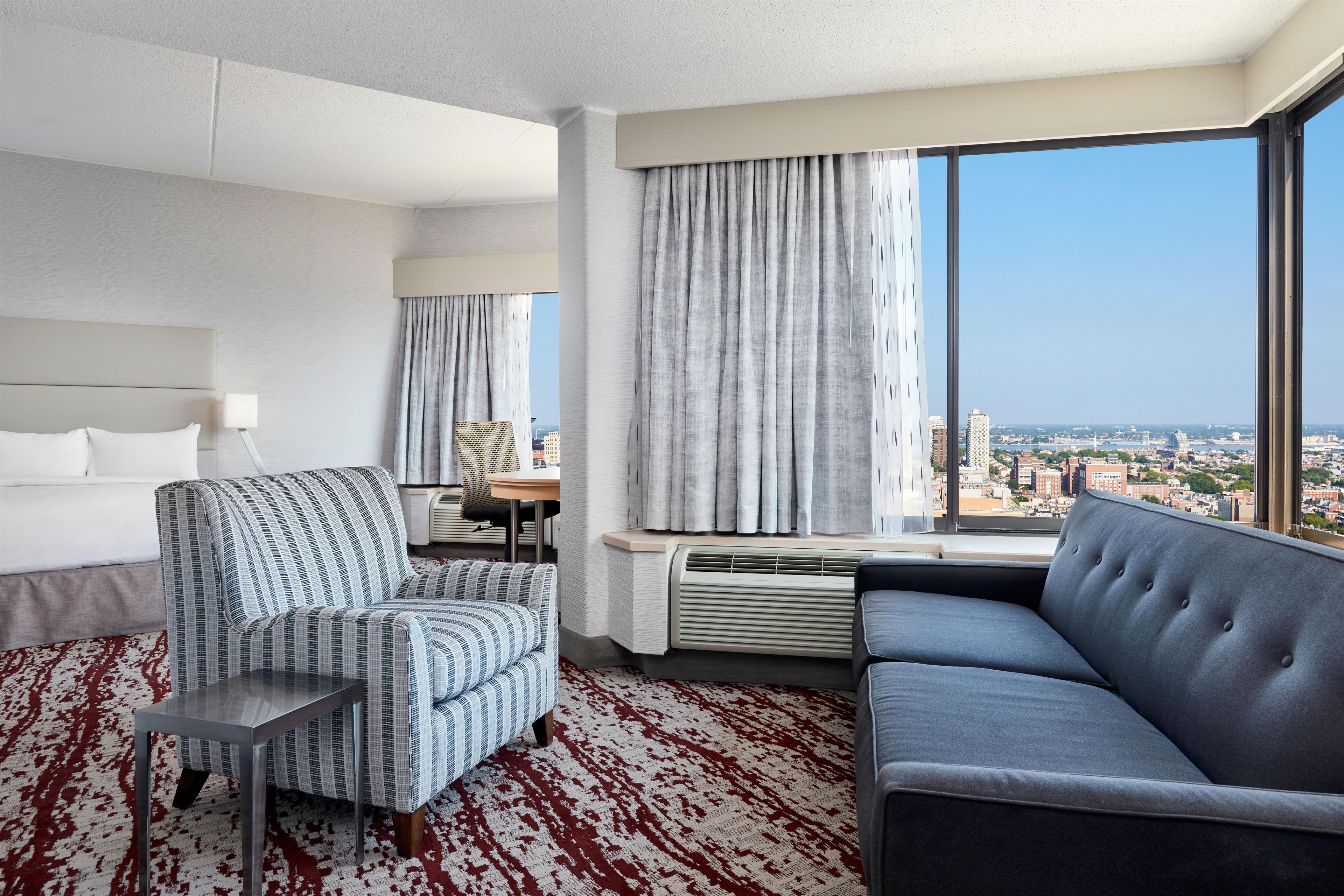 A hotel room with a large window, a sofa, a chair, a bed, and a desk.
