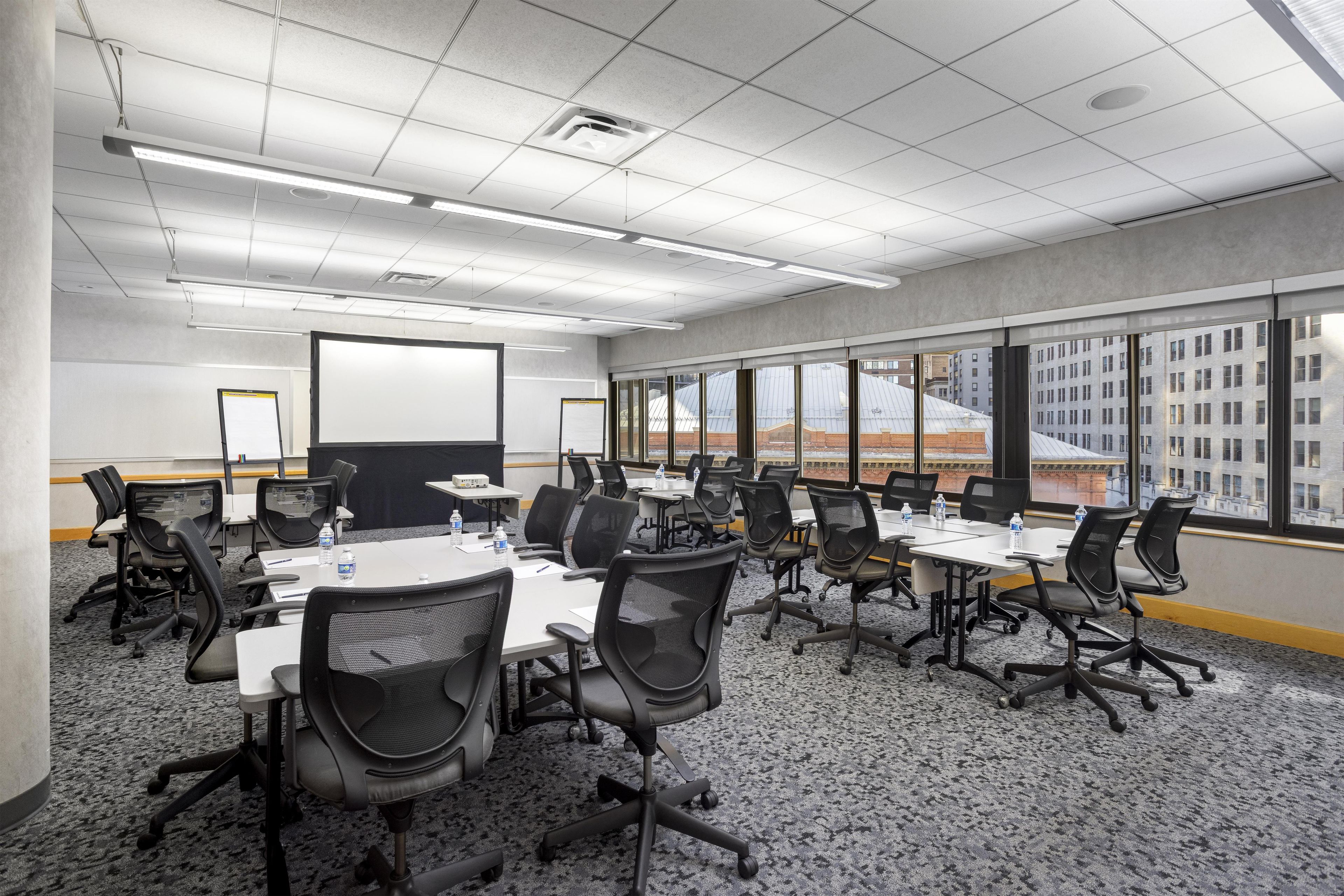 Un salón de conferencias con varias mesas y sillas, amplias ventanas que dan a vistas de edificios y una pantalla de proyector.