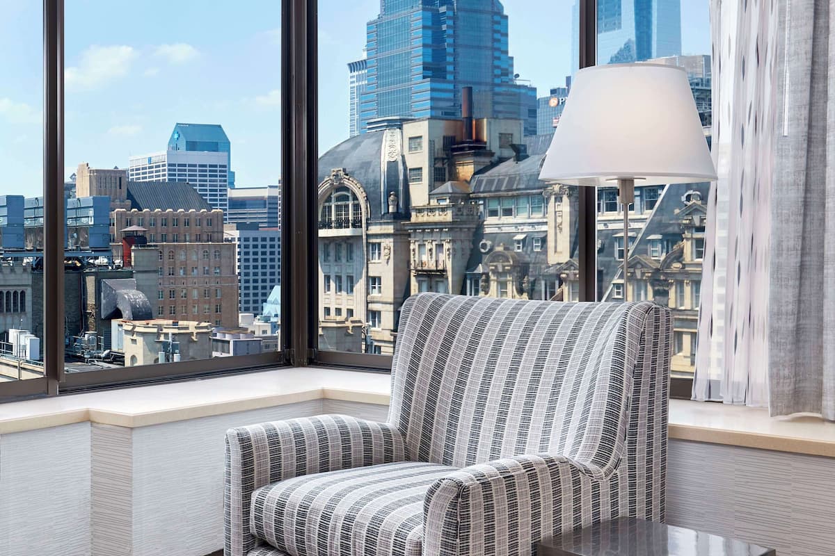 A modern room with a large window offering a city view, a striped armchair, and a floor lamp.