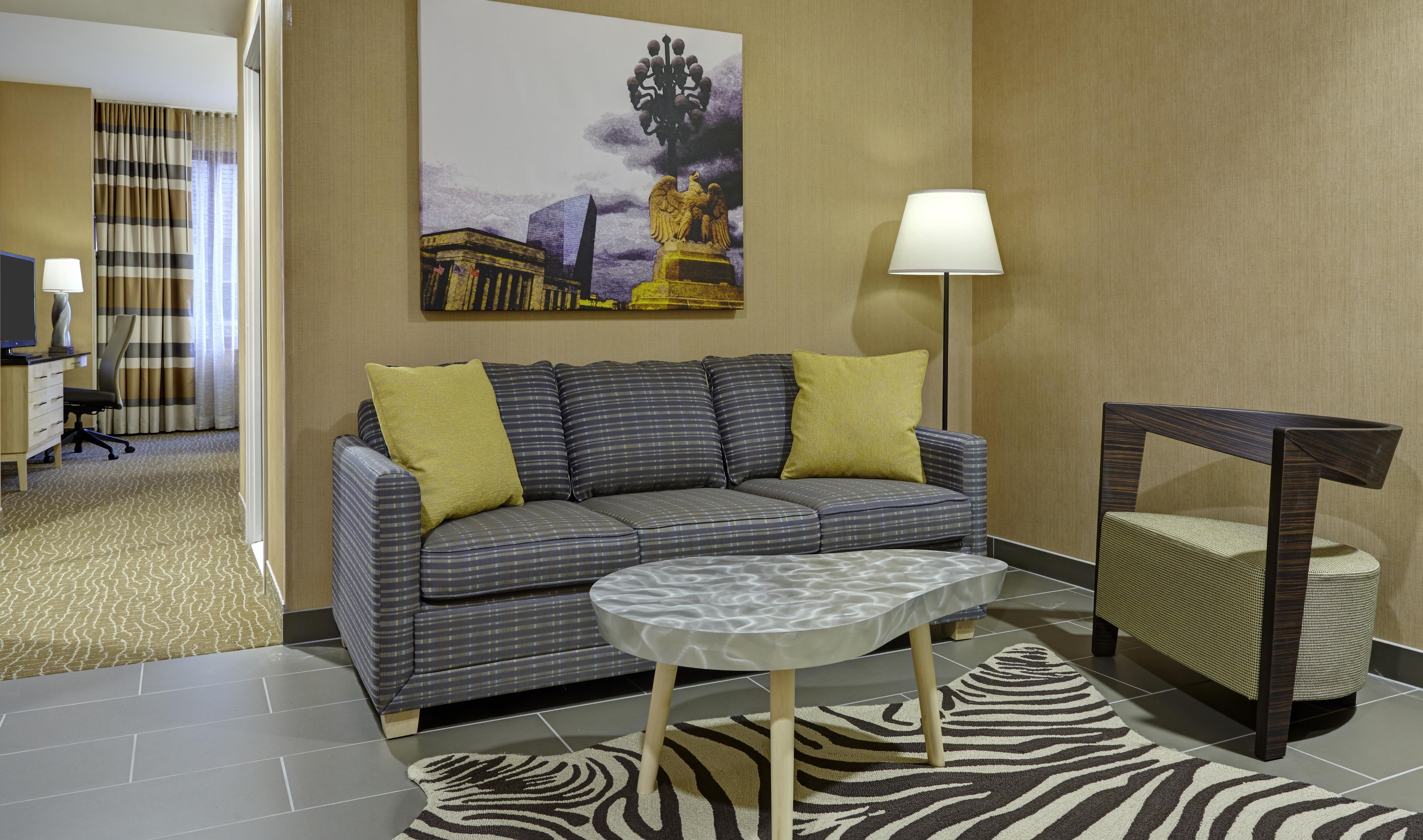 Une chambre d’hôtel avec un canapé, une petite table ronde, une lampe, une chaise et un tapis à motifs zébrés.