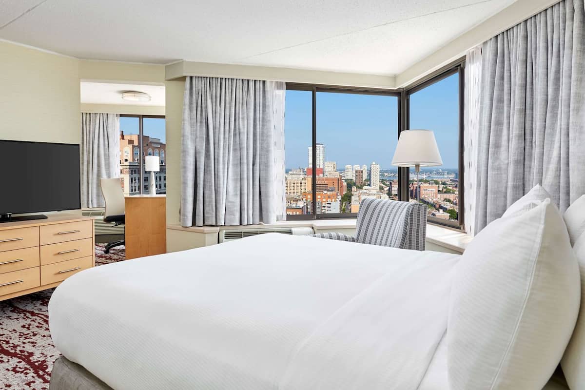 A hotel room with a large bed, a TV, a desk with a lamp, and a view of the cityscape.