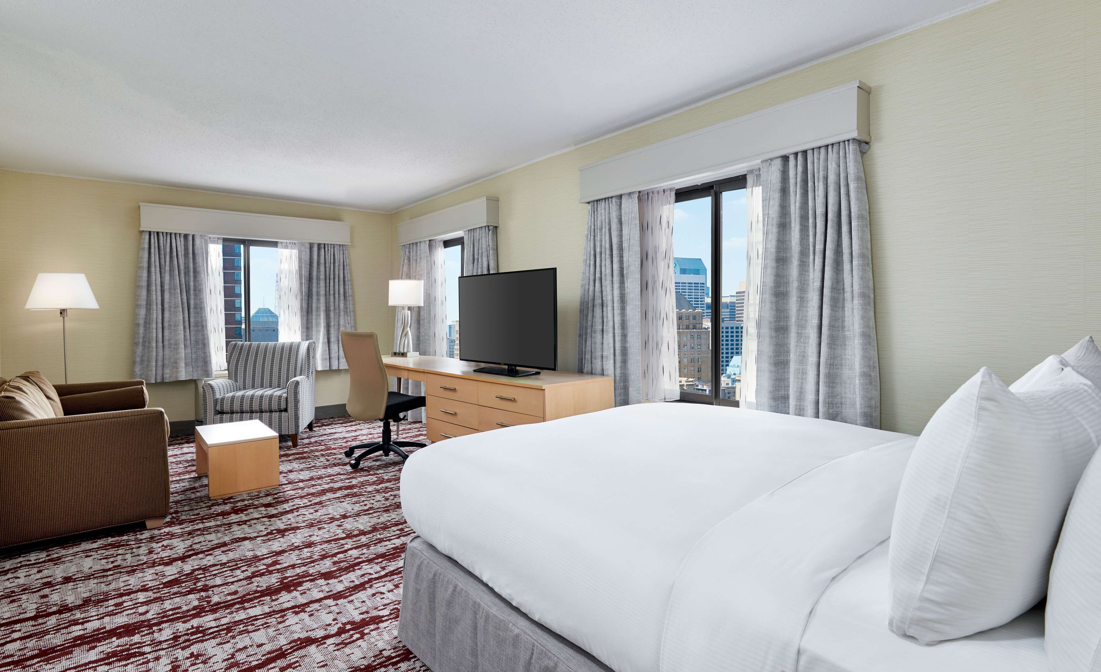 Una habitación de hotel con cama, escritorio, silla, televisor y ventanas amplias con cortinas.