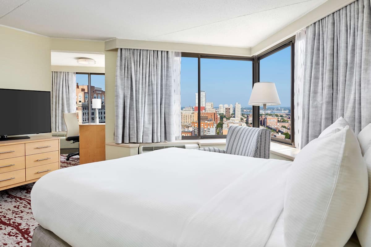 A hotel room with a large bed, a TV, a desk with a lamp, and a view of the cityscape through large windows.