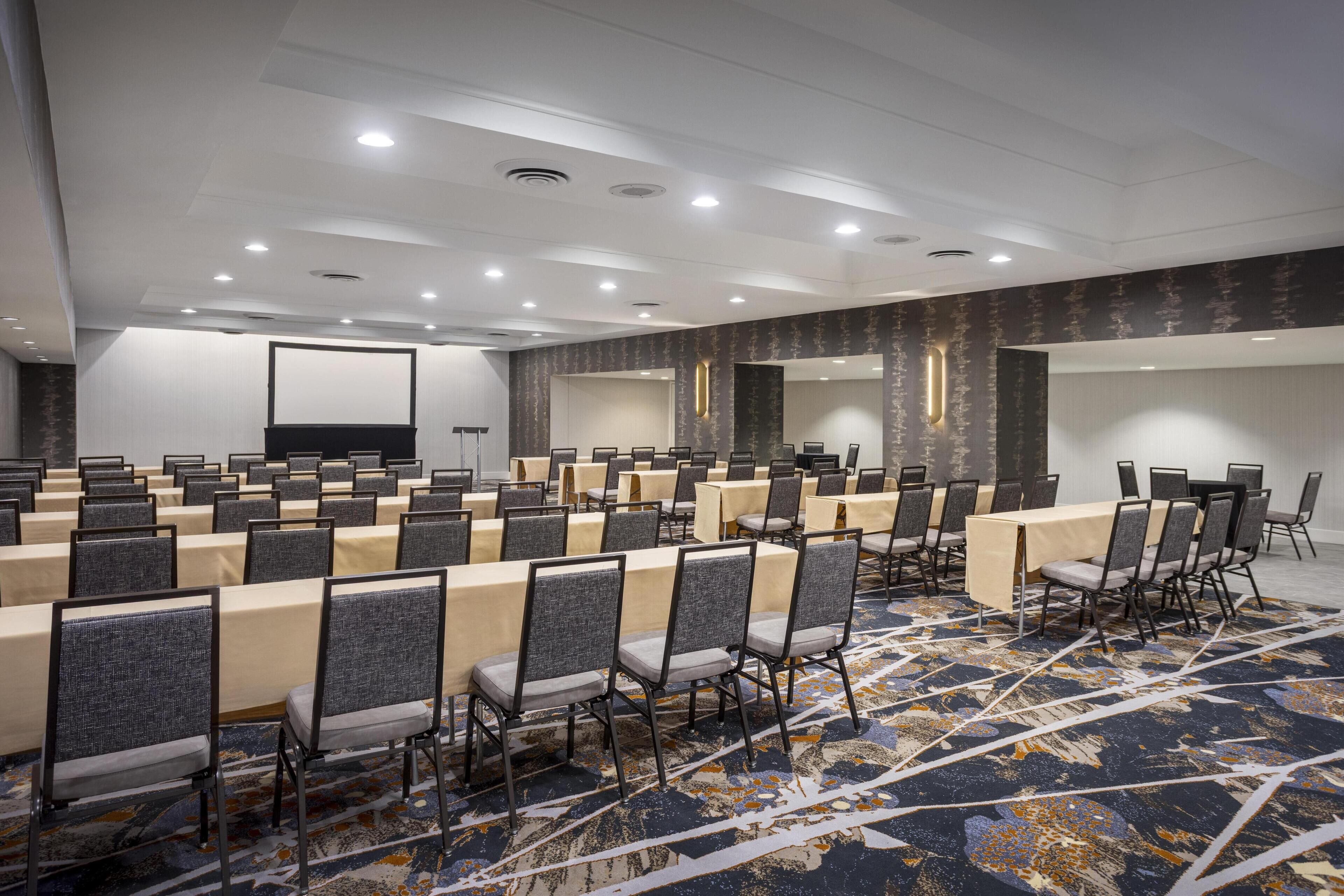 A conference room with rows of tables and chairs, a large screen, and a patterned carpet.