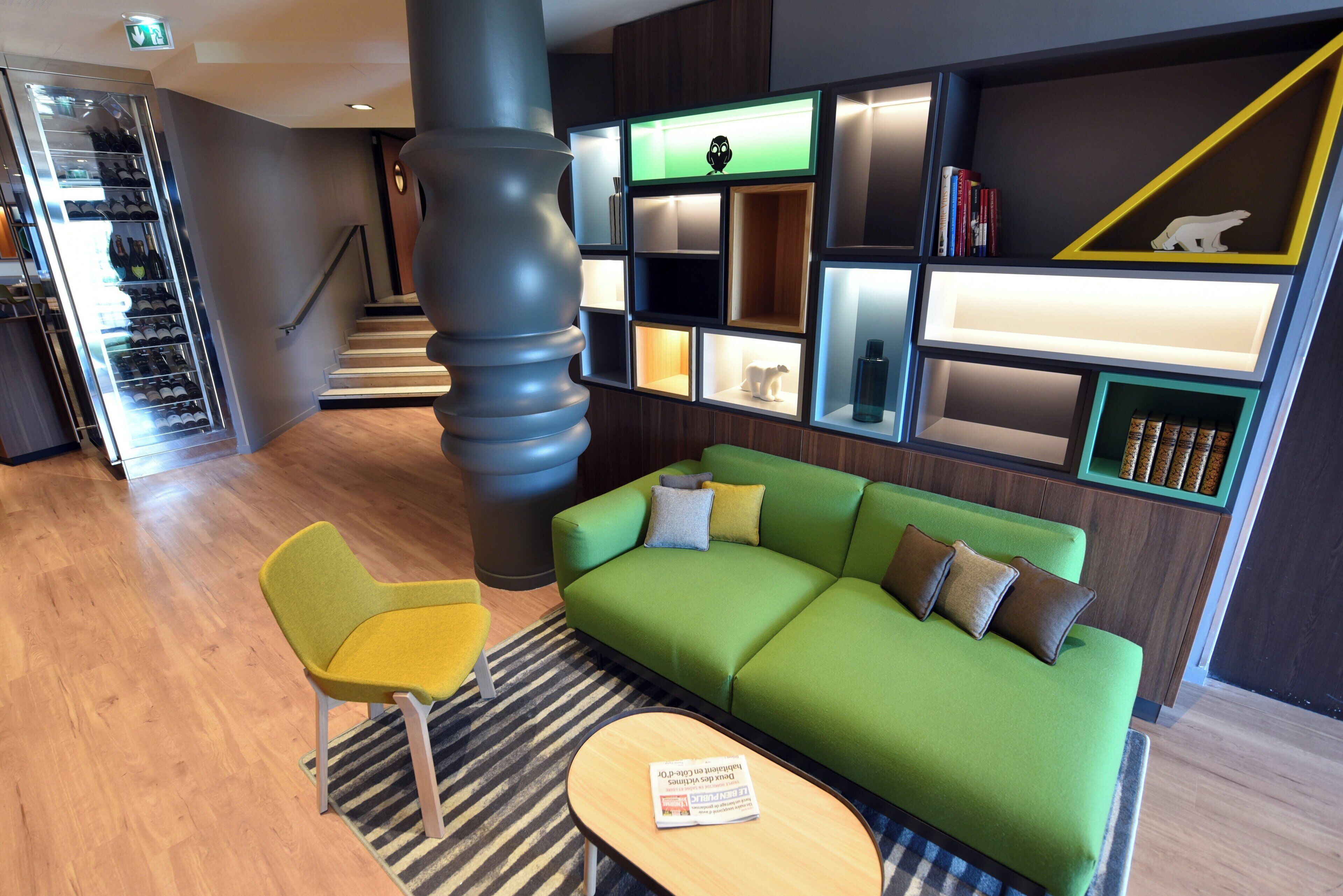 A modern interior with a green sofa, a yellow chair, a striped rug, and a bookshelf with various items.