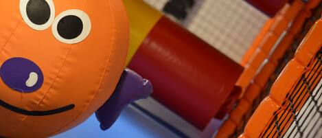 A plush toy with a smiling face and large eyes, placed near a play structure with a blue floor and orange netting.