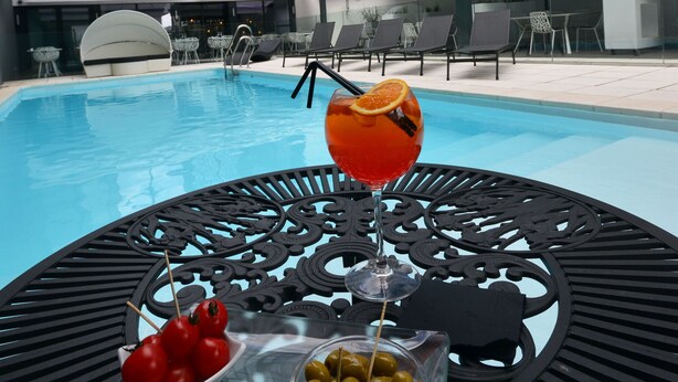 Une table placée au bord de la piscine, avec une boisson, des olives et des tomates, entourée de chaises longues et d’un bâtiment moderne doté de grandes fenêtres.