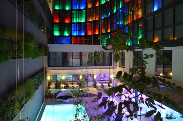 A modern hotel interior with a pool area, indoor plants, and a dining space.
