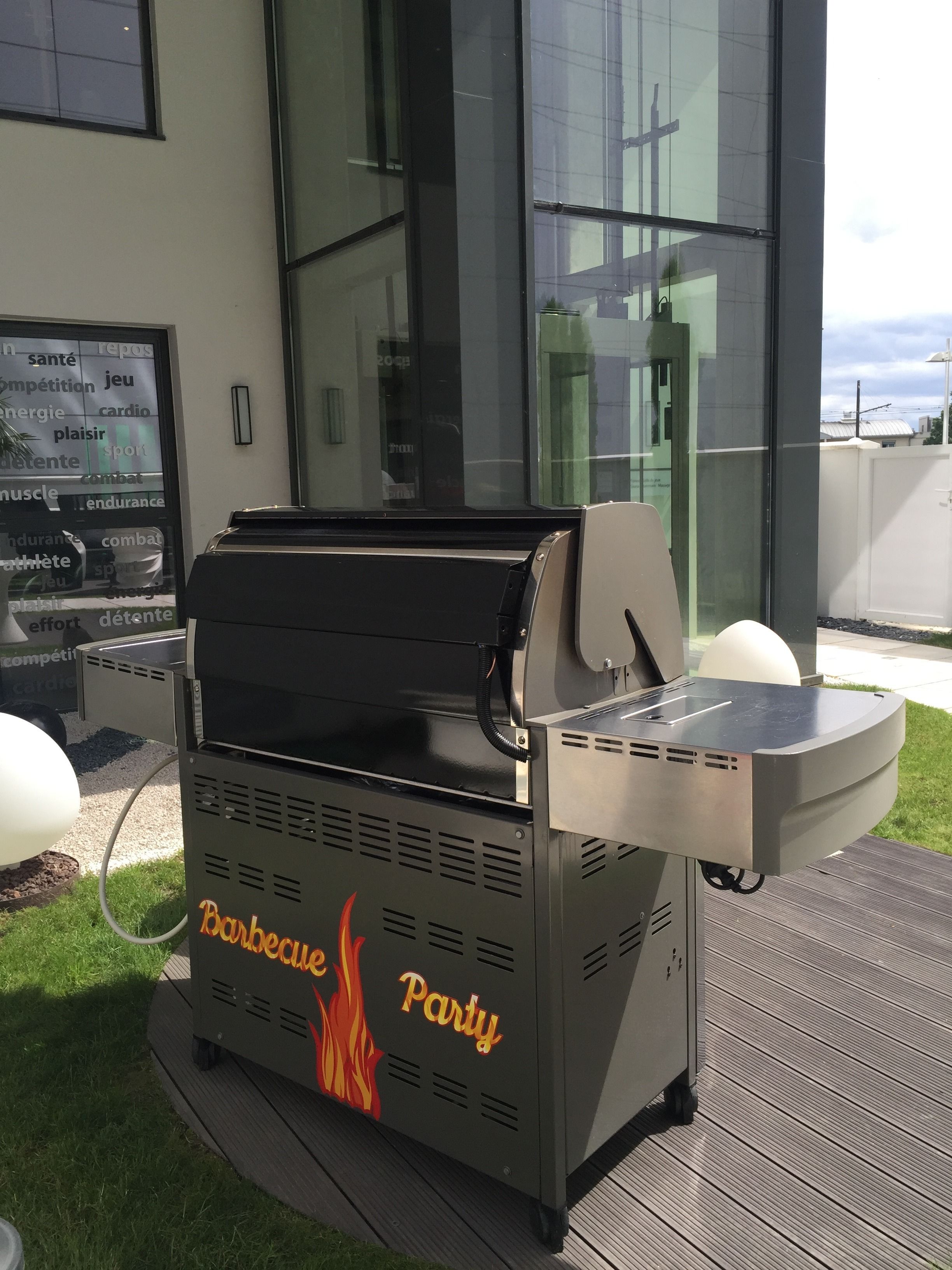 A barbecue grill with 'Barbecue Party' branding, located outdoors on a patio with a modern building in the background.