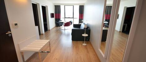 Una habitación de hotel moderna con suelo de madera, un sofá negro, una silla roja, una mesita y una lámpara.