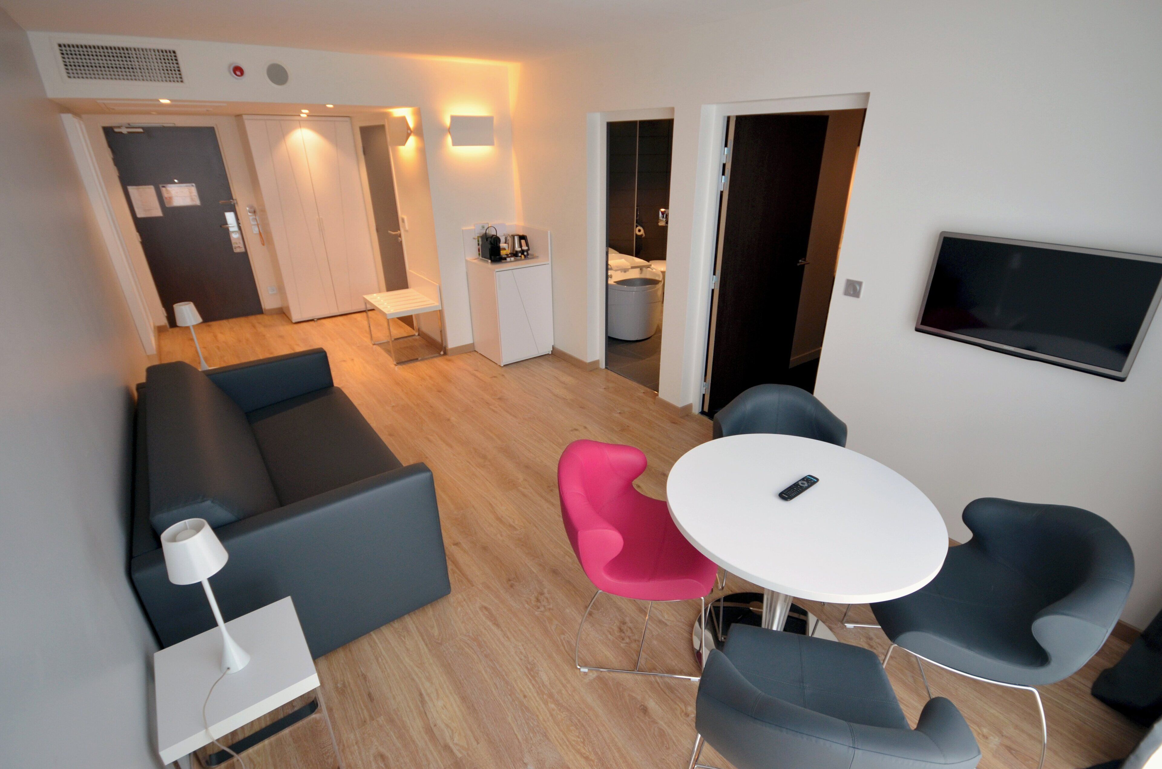 A modern living area with a sofa, a small table, and two chairs. There is a television mounted on the wall, and a kitchenette is visible in the background.