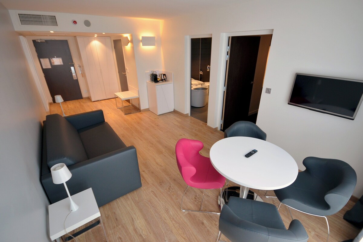 A modern living room with a sofa, a pink chair, a round table, and a TV mounted on the wall.