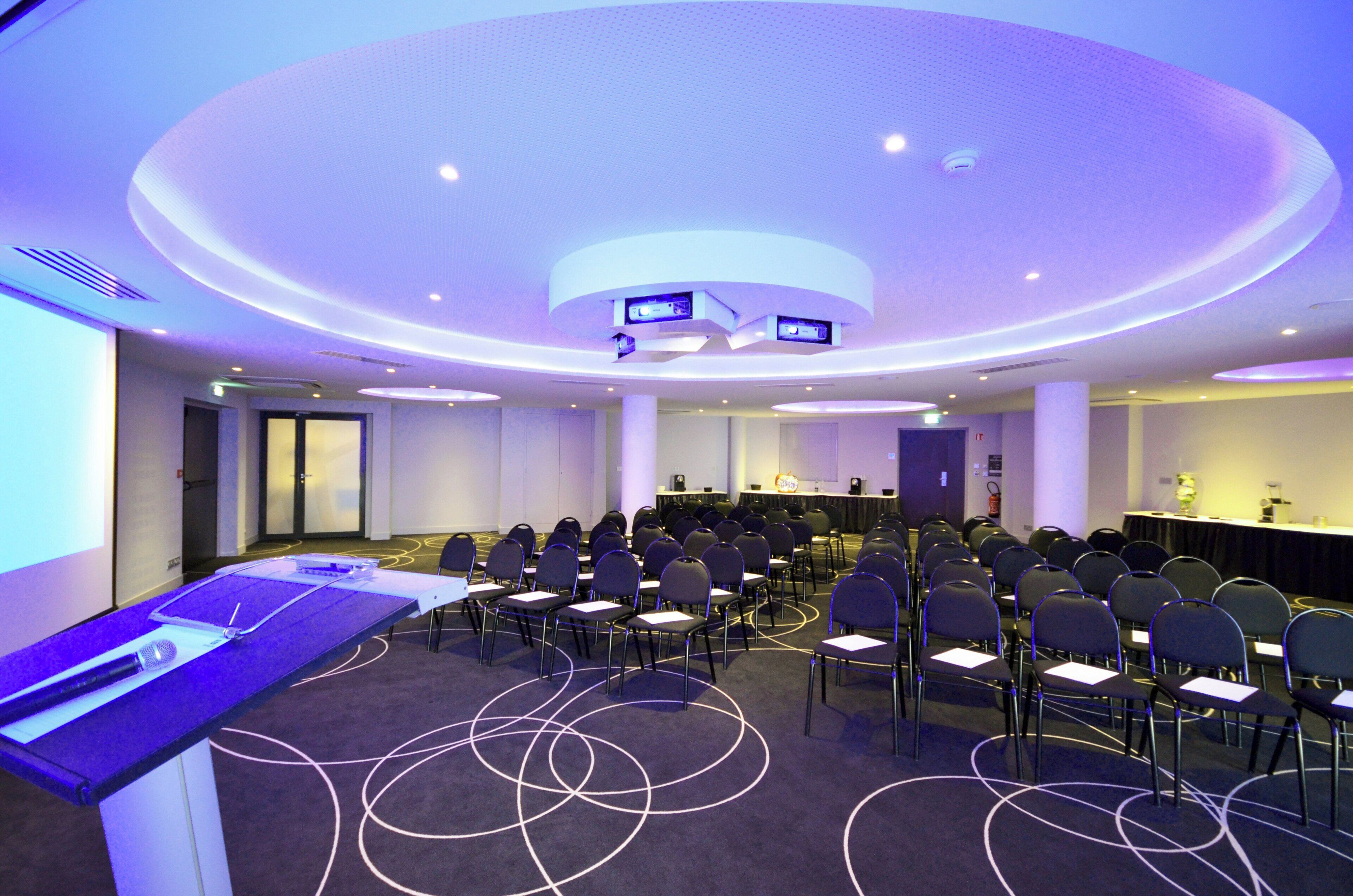 Un salón de conferencias con filas de sillas orientadas hacia una zona de presentaciones, equipado con un atril y una pantalla.