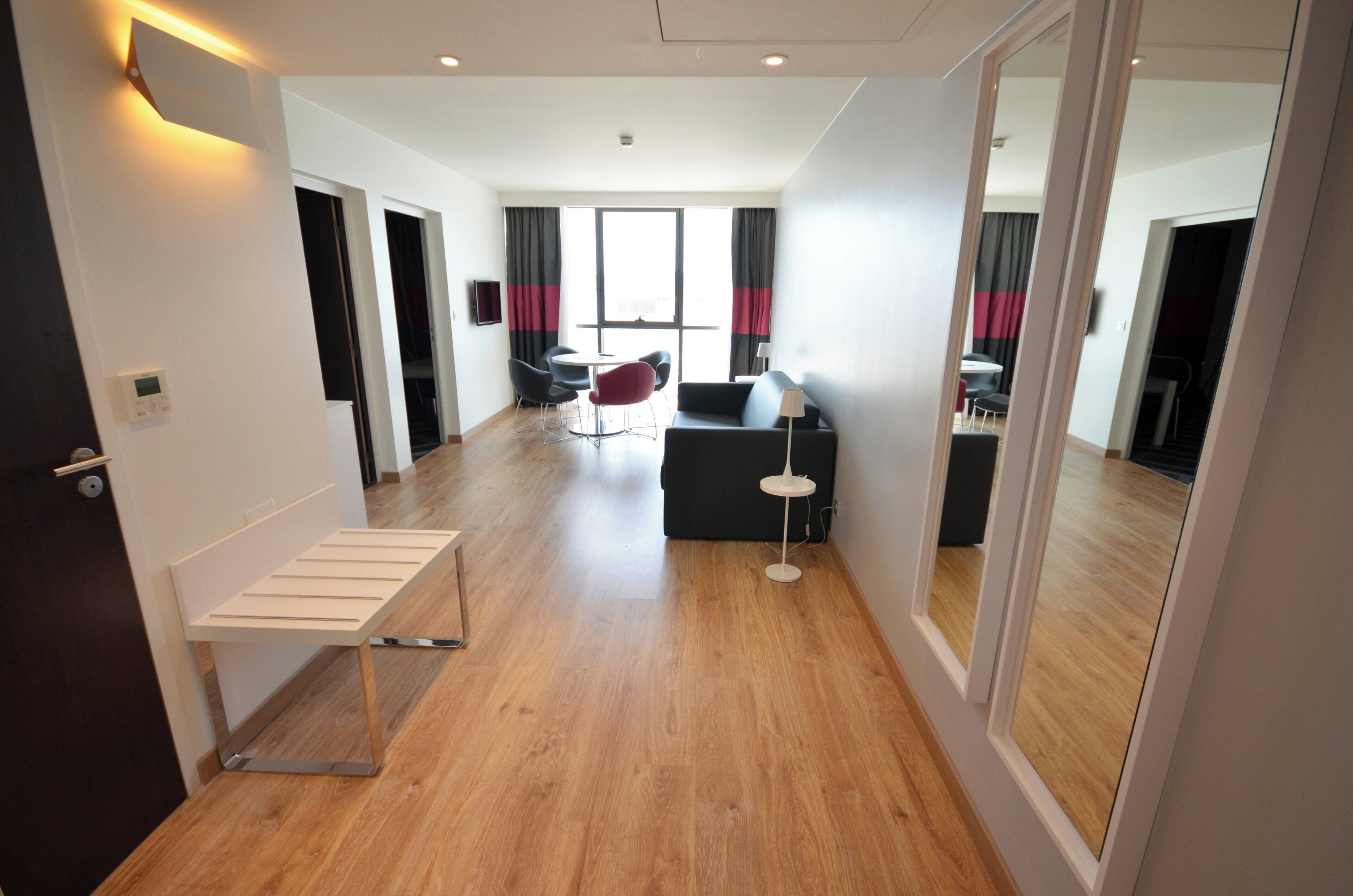 Ein modernes Hotelzimmer mit Holzboden, einer Sitzecke mit Sofa und Sesseln, einem kleinen Tisch und einem an der Wand befestigten Fernseher.