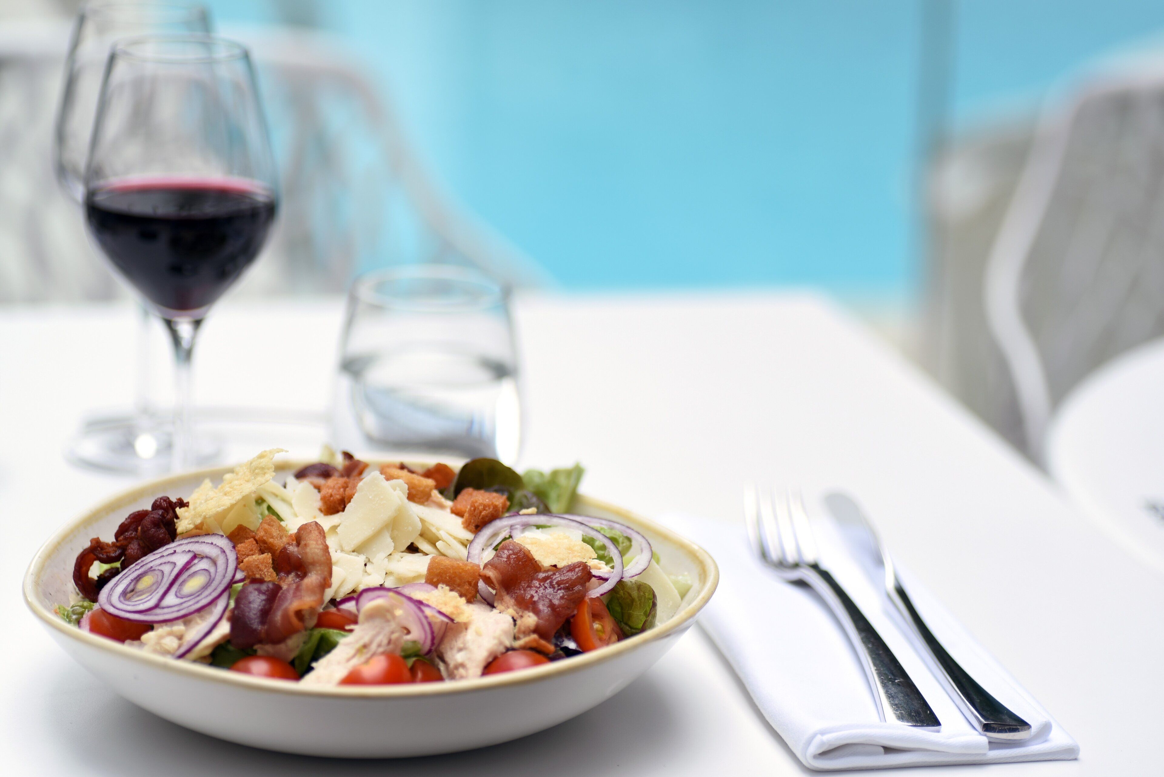 A bowl of salad with croutons, cheese, and bacon, accompanied by a glass of red wine and a water glass on a white table.