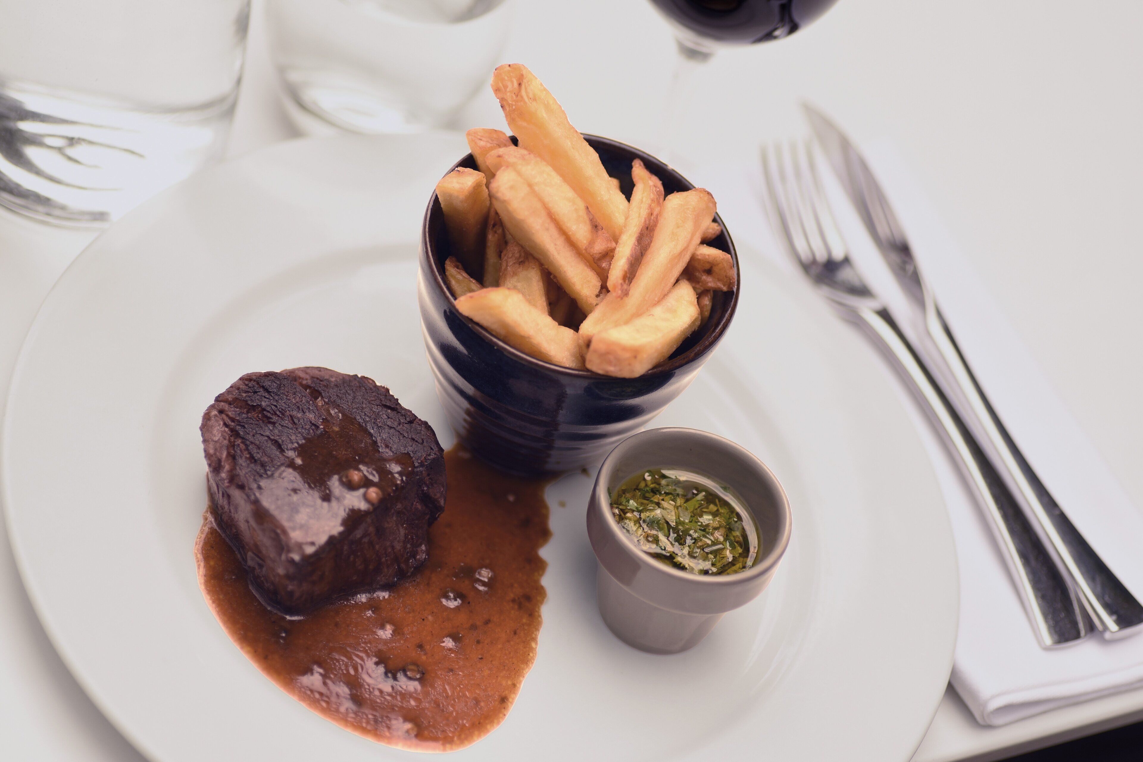 A plate with a steak, fries, and a sauce, accompanied by a small bowl of green sauce.