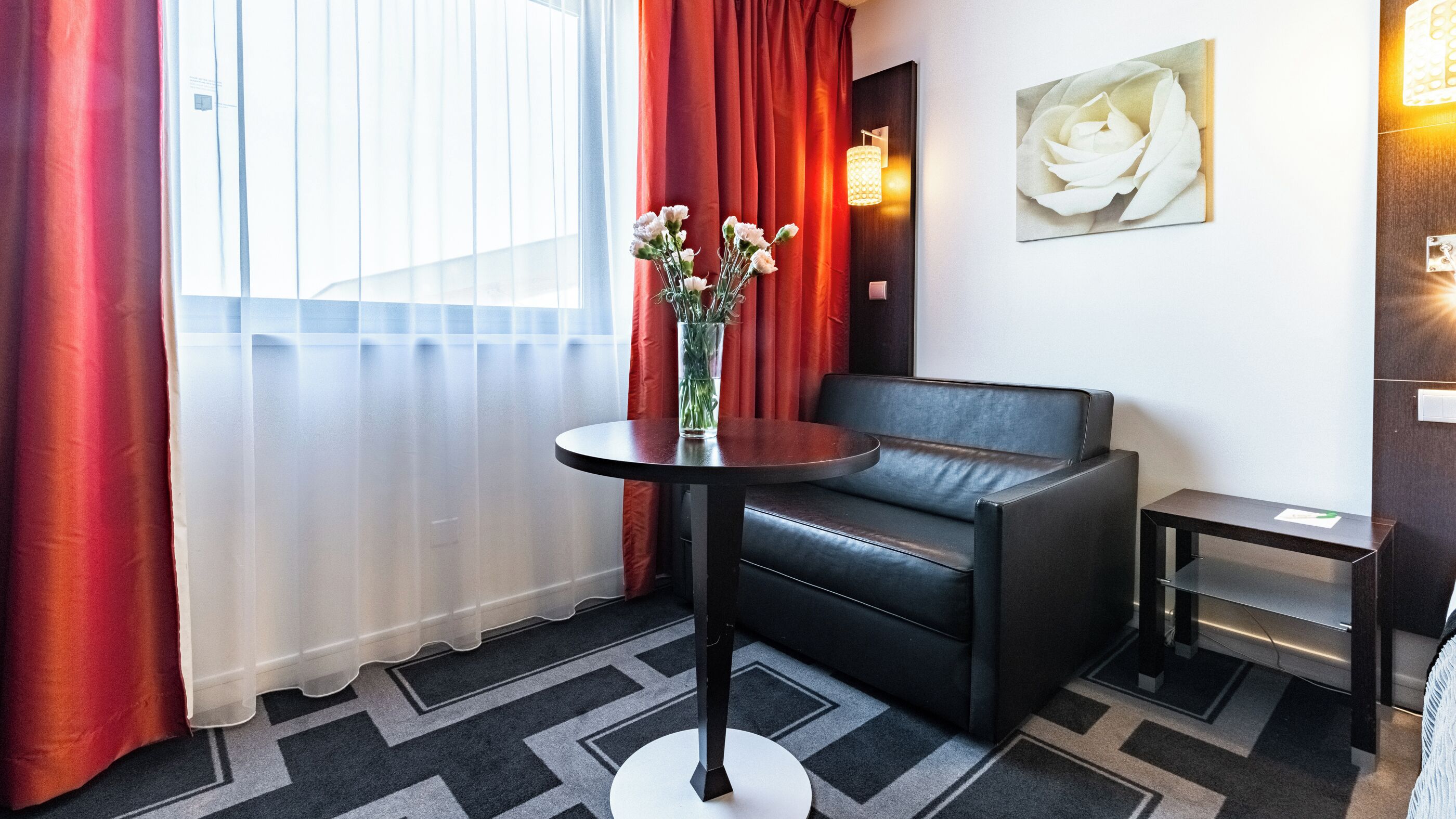 A modern hotel room with a black leather sofa, a round table with a vase of flowers, a painting of a rose on the wall, and a small side table.