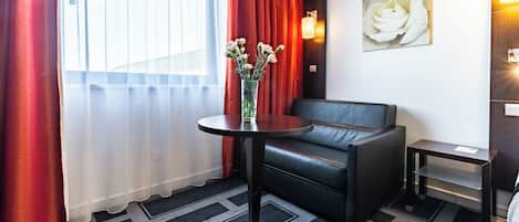 A modern hotel room with a black leather sofa, a round table with a vase of flowers, a painting of a rose on the wall, and a small side table.