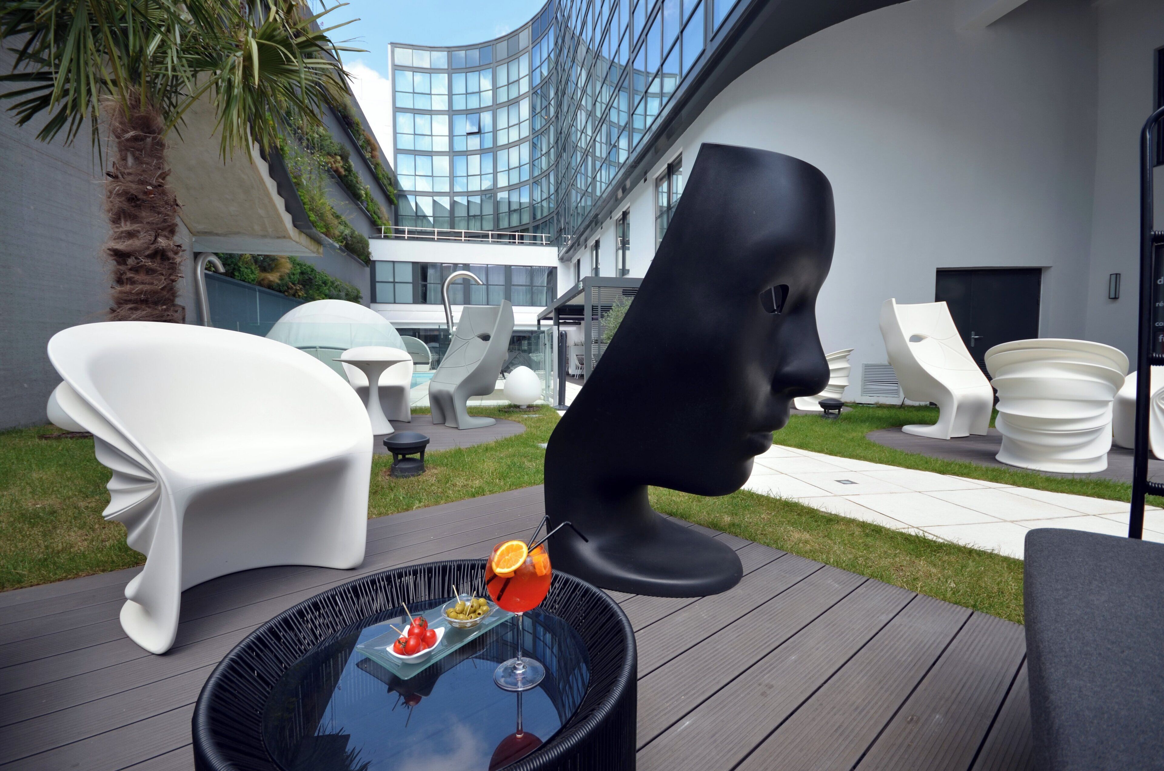 A modern outdoor seating area with unique white and black chairs, a glass table with a drink and olives, and a view of a contemporary building with a curved glass facade.