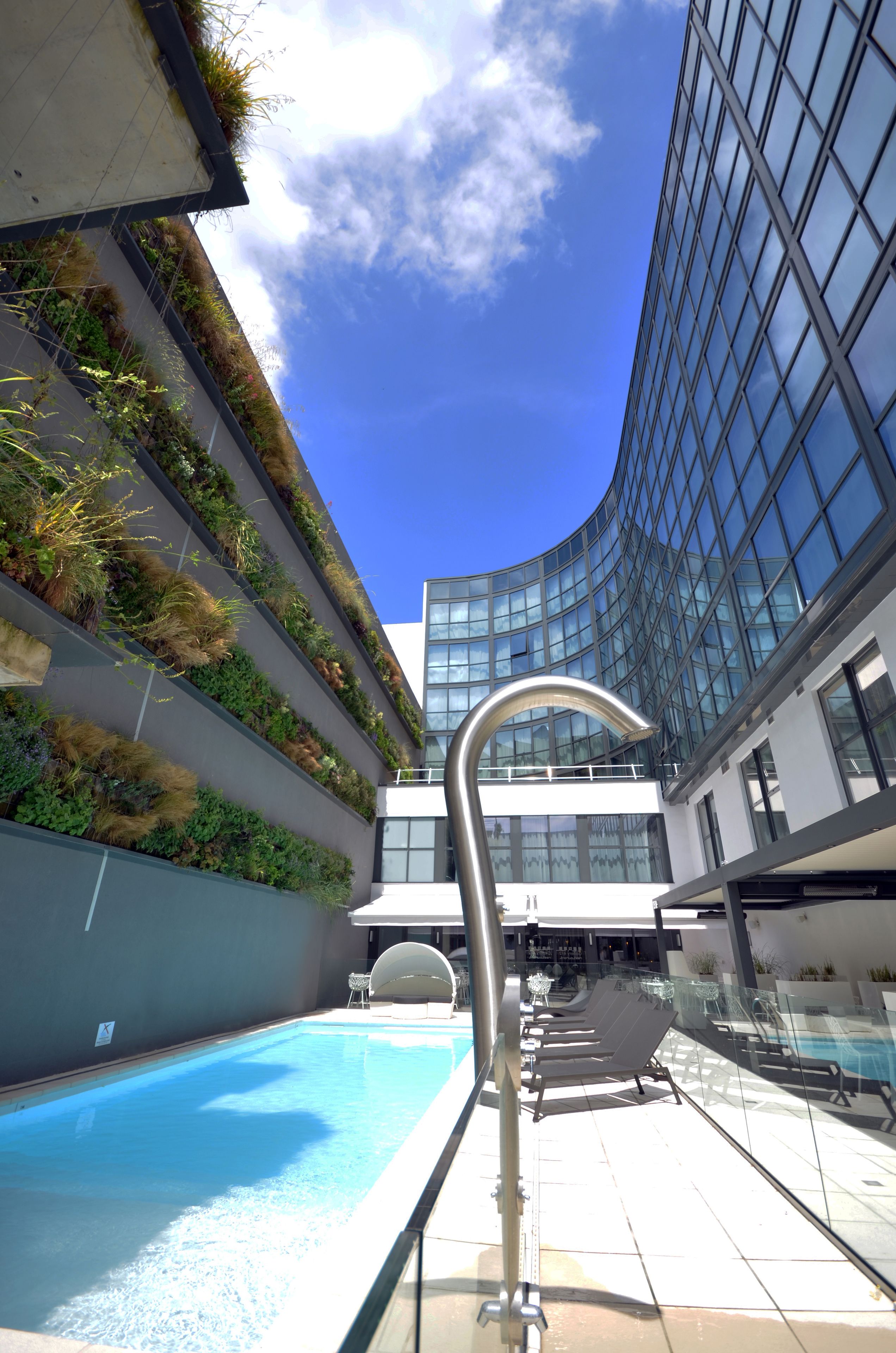 Ein modernes Gebäude mit einer geschwungenen Glasfassade, einem Pool auf dem Dach mit Liegestühlen und einer vertikalen Begrünung.
