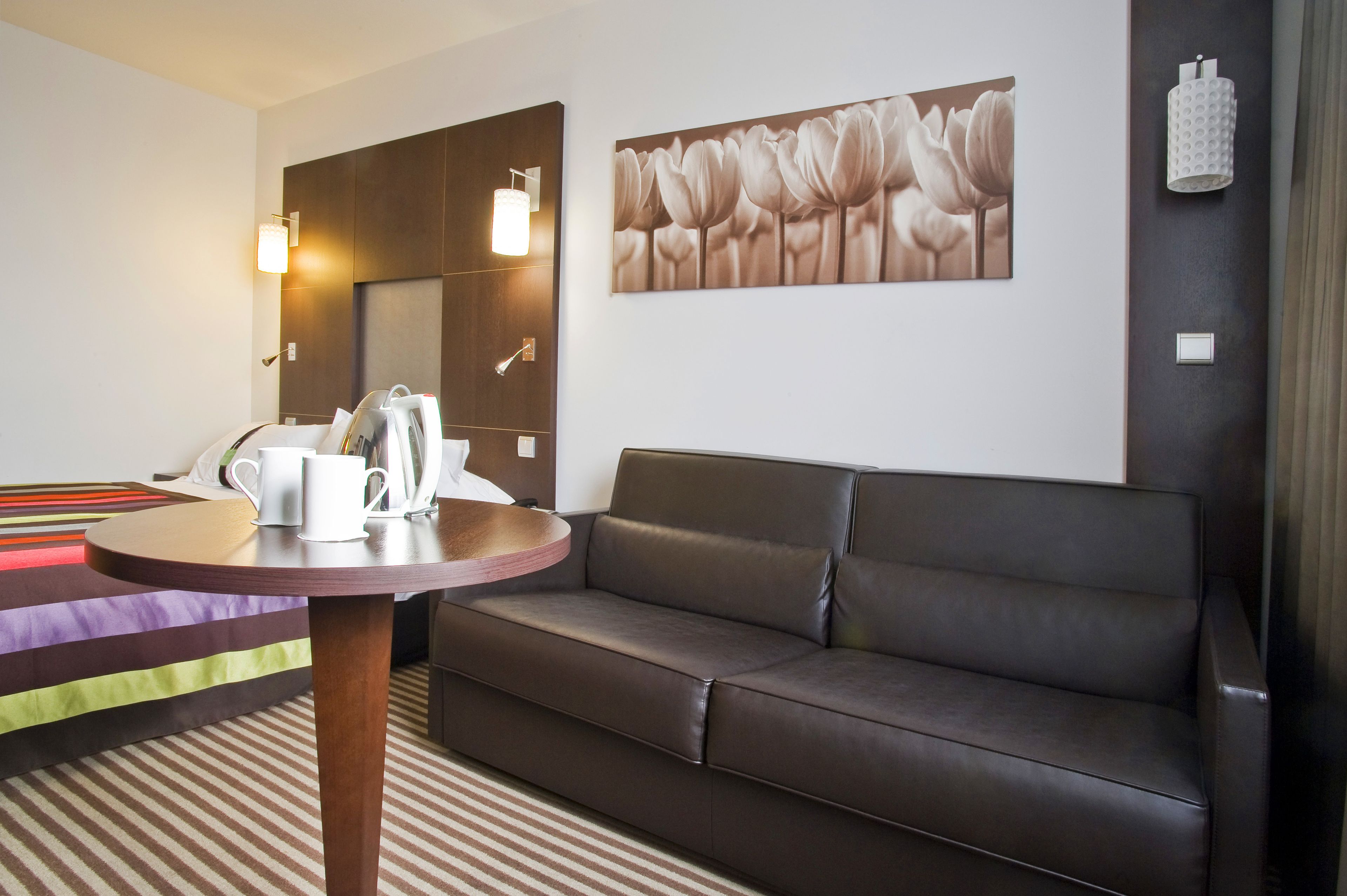 A modern hotel room with a dark leather sofa, a round wooden table with a white mug and a small white lamp, and a large floral artwork on the wall.
