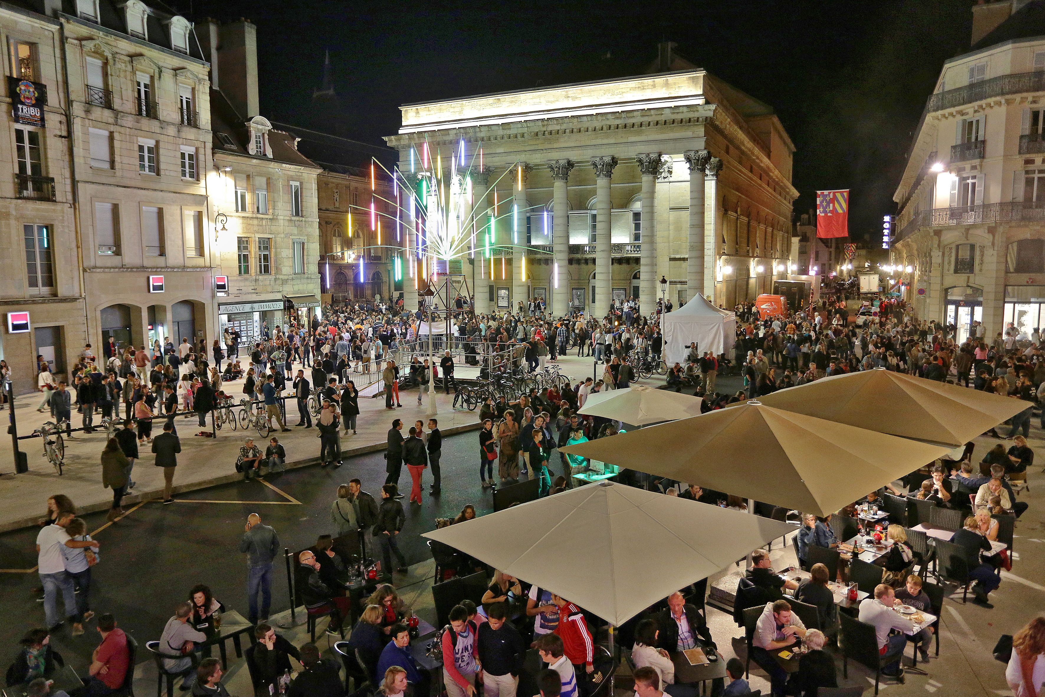 A crowded square with outdoor seating and a large building in the background.