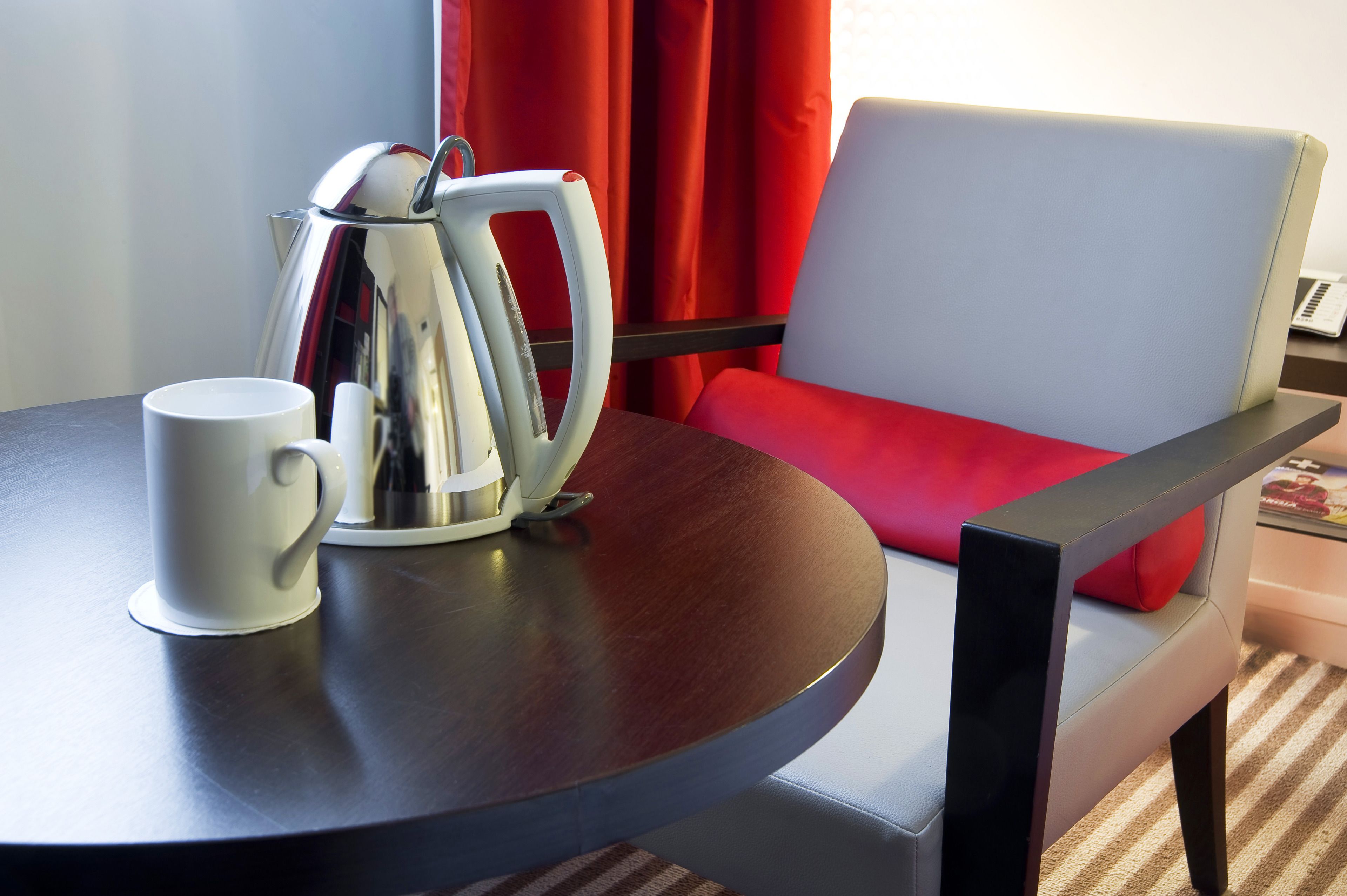 A table with a white mug and a silver electric kettle, a red chair with a white cushion, and a laptop on a small table.