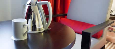 A table with a white mug and a silver electric kettle, a red chair with a white cushion, and a laptop on a small table.