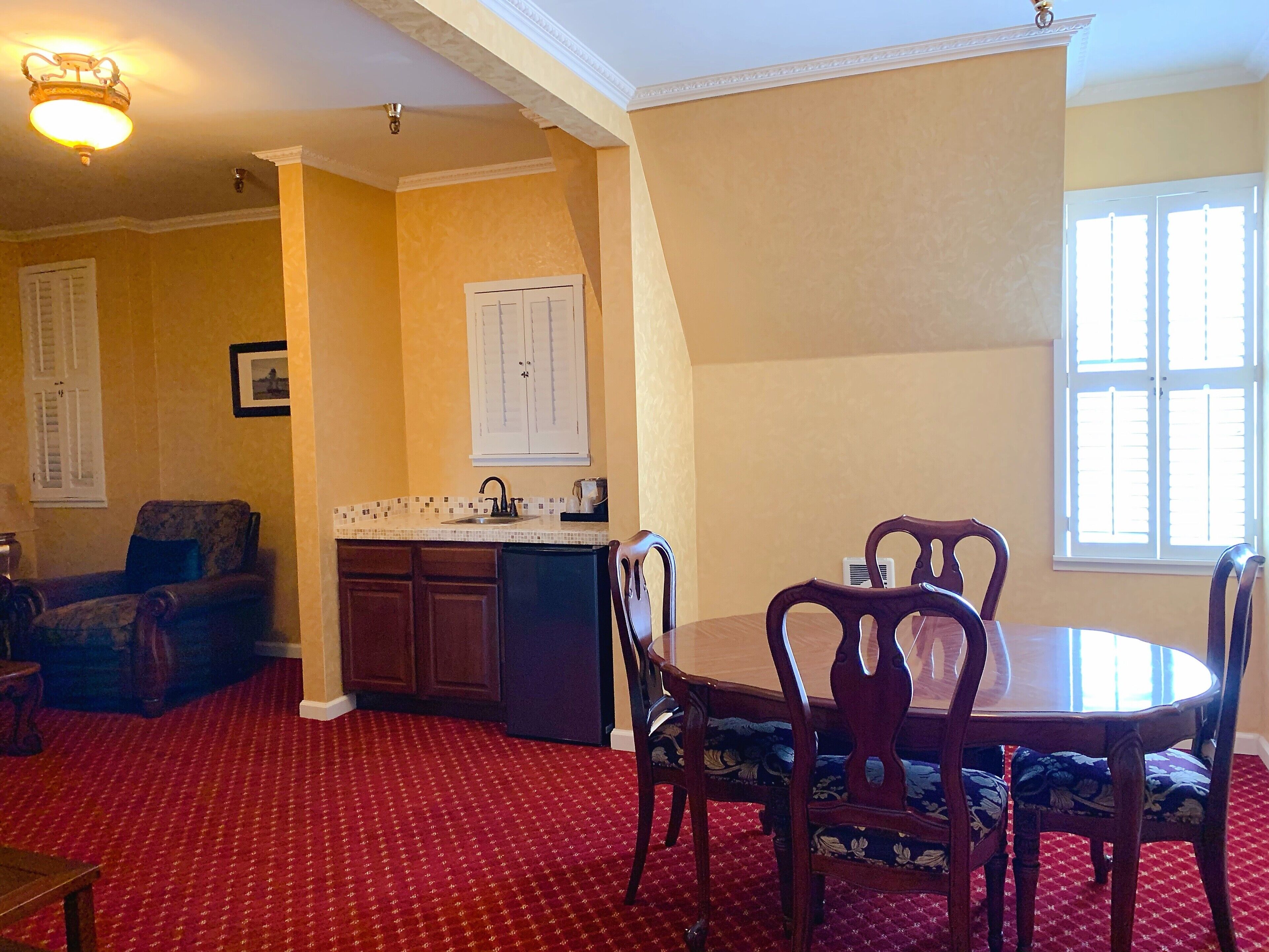 A dining area with a round table and chairs, a kitchen with a tiled backsplash, and a red patterned carpet.