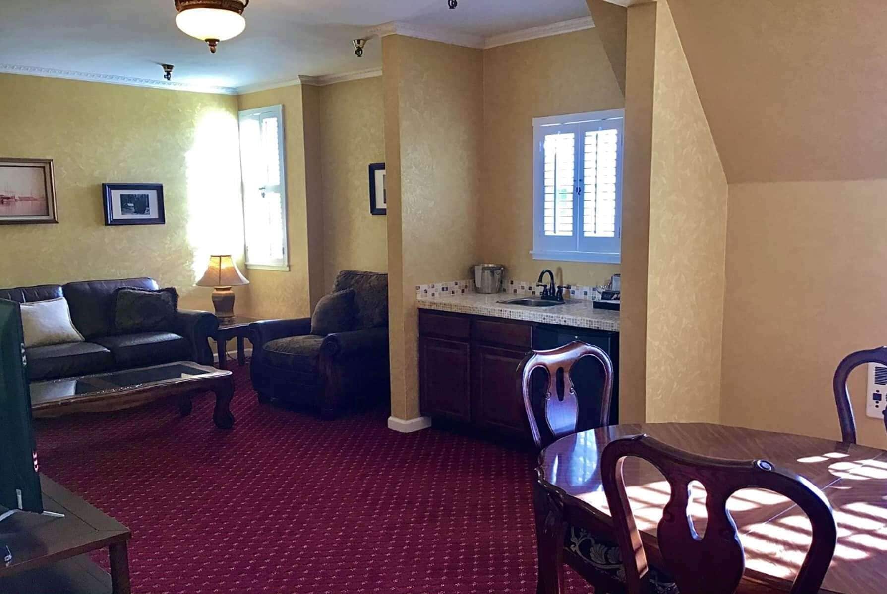 A living room with a sofa, armchair, and a dining table with chairs. There is a kitchen area with a window and a sink.