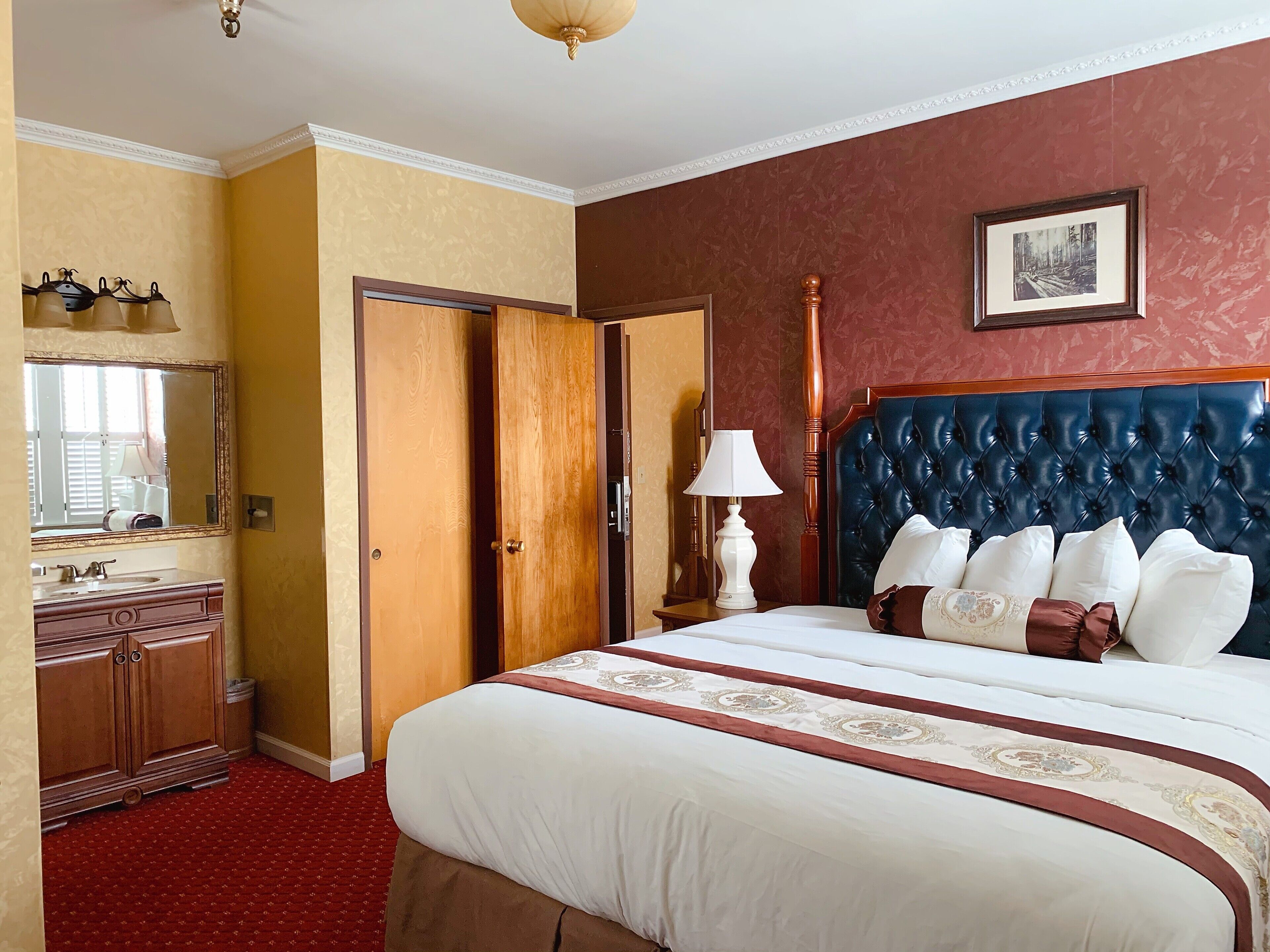 A hotel room with a large bed, a wooden wardrobe, a mirror, a bedside table with a lamp, and a framed picture on the wall.